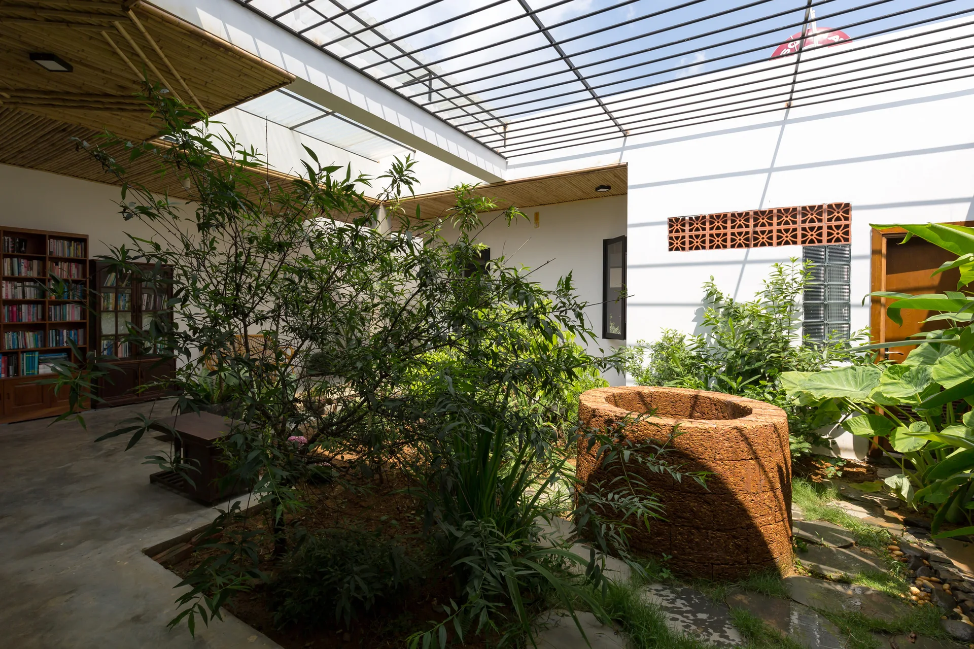 A bright indoor-outdoor courtyard features a central garden with lush green plants, a rustic stone well, a bamboo ceiling, and white walls.