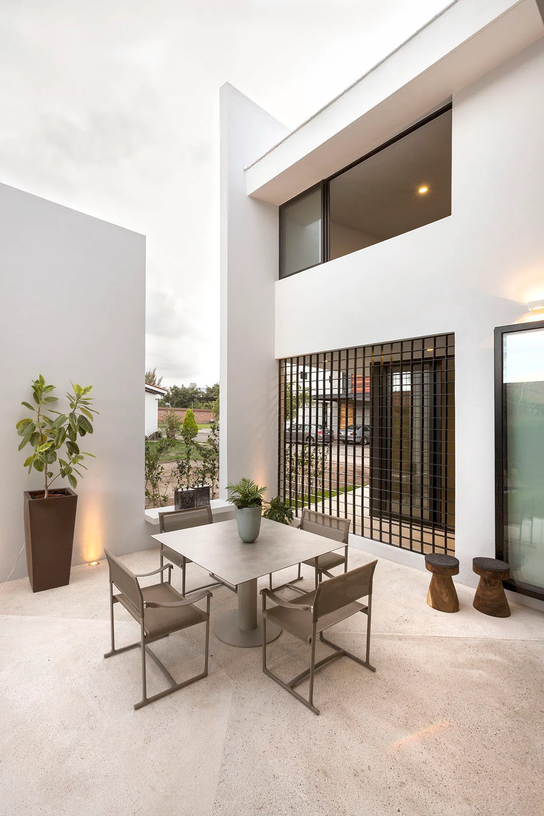 A modern outdoor patio features a square grey dining table and four chairs on light stone tile, set against clean white minimalist exterior walls.