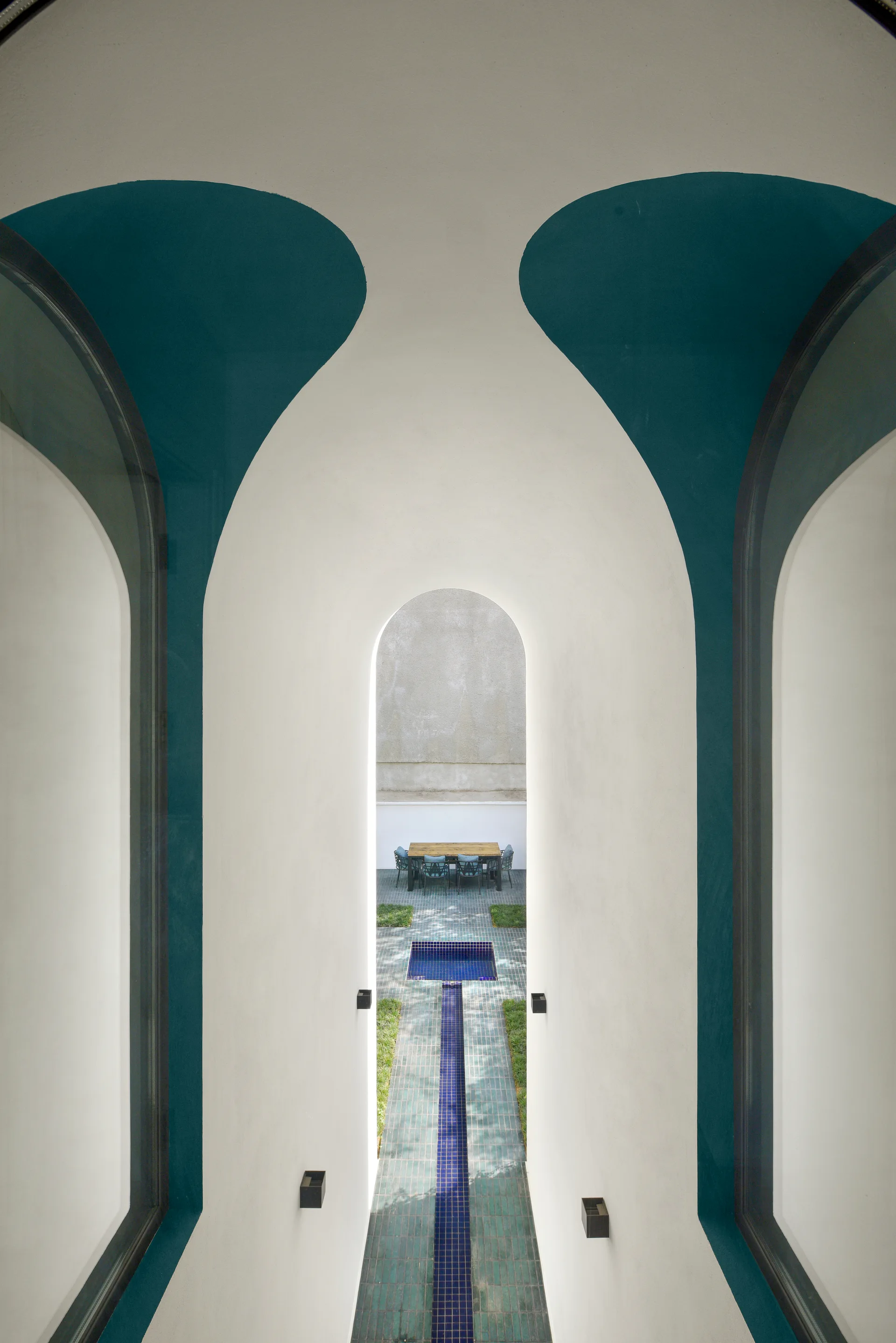Symmetrical white stucco archways trimmed in teal frame a courtyard view of a tiled walkway, a small blue pool, and an outdoor dining set.