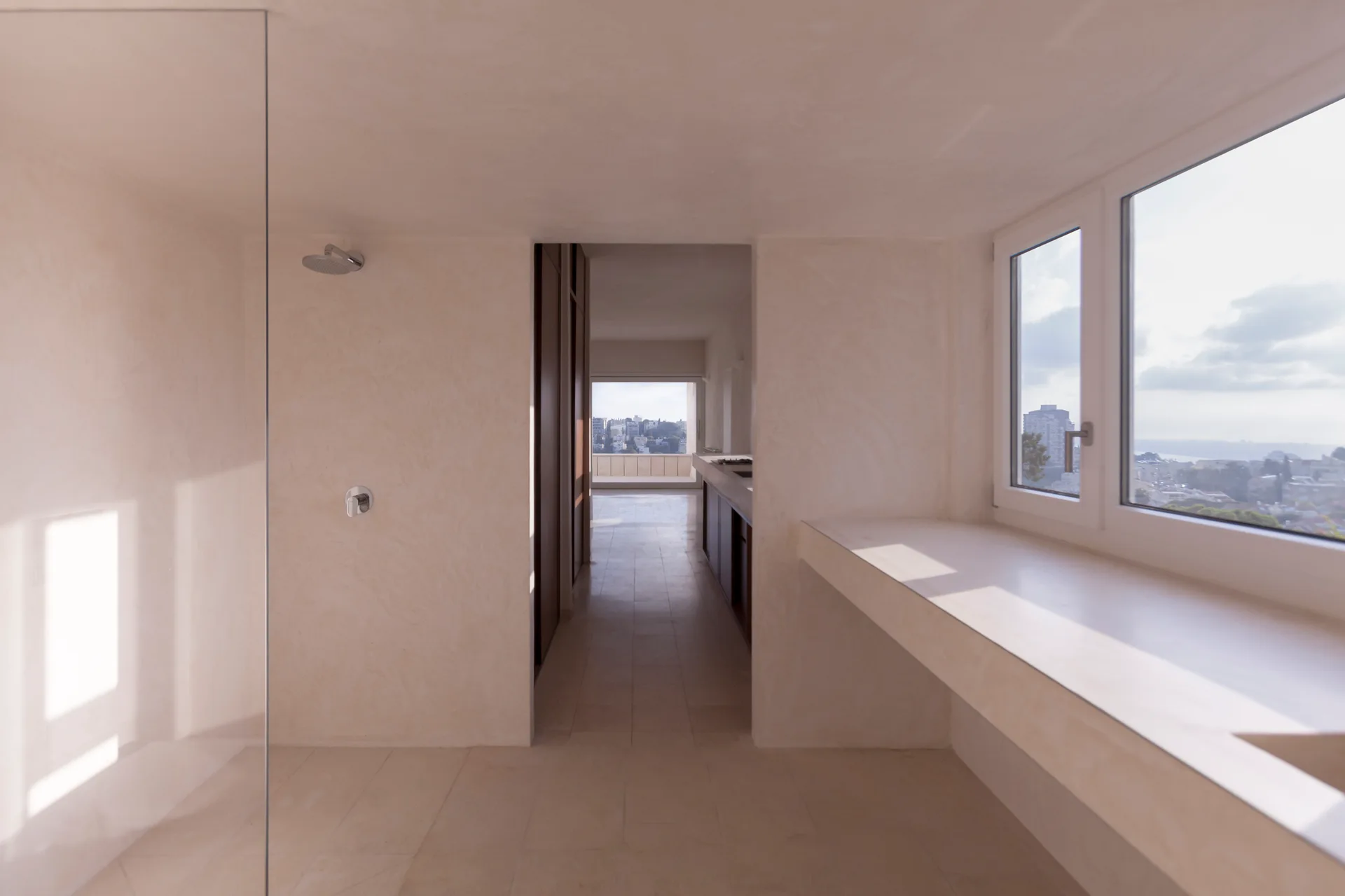A minimalist bathroom with beige plaster walls, a stone tile floor, a glass shower partition, and a built-in counter beneath a window.