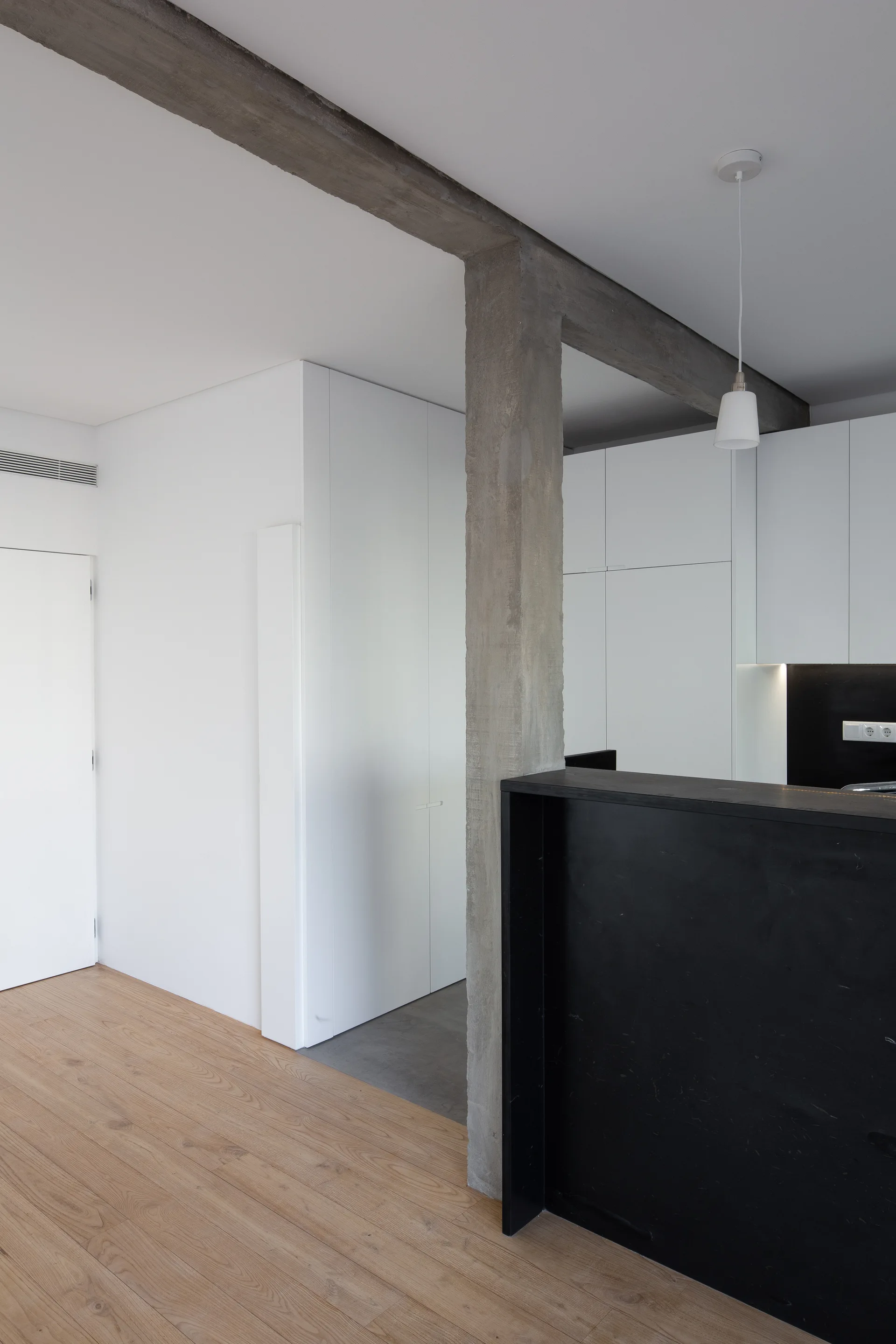 A minimalist kitchen featuring white handleless cabinets, a matte black island, light wood flooring, and an exposed raw concrete column.