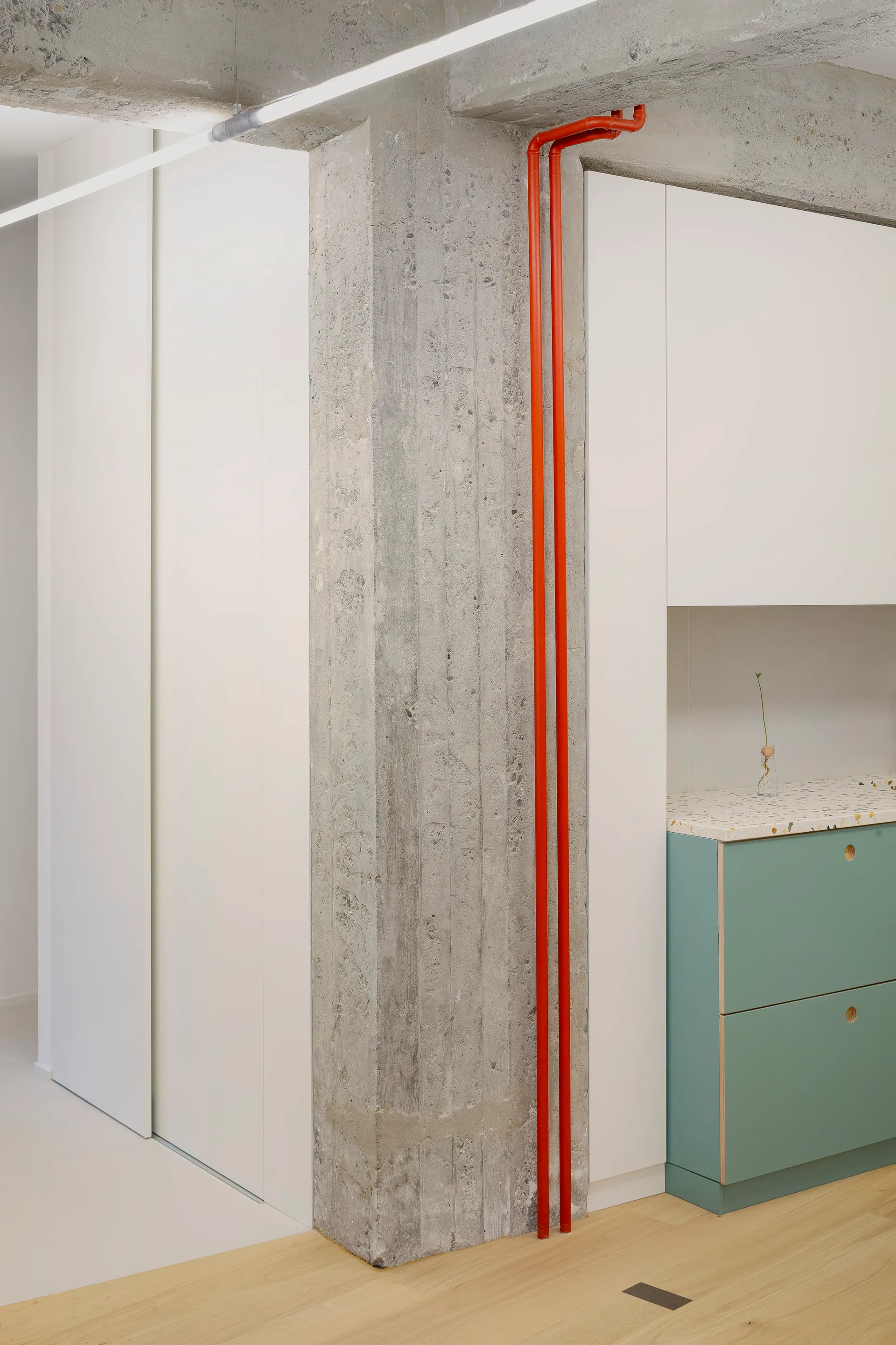 An exposed concrete pillar with two bright red pipes stands next to minimalist white and green kitchen cabinets with a terrazzo countertop.