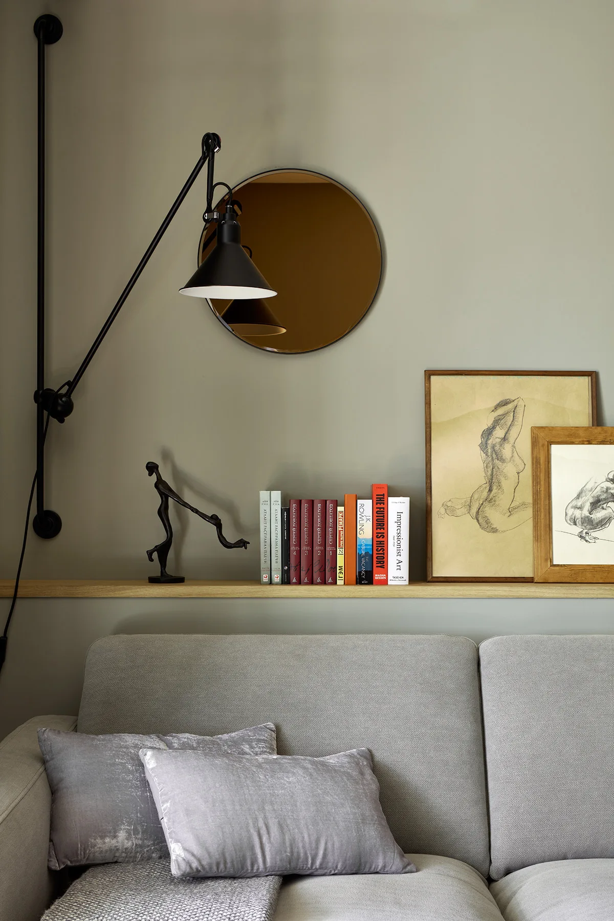 A light grey herringbone sofa sits beneath a slim wooden ledge displaying books and framed sketches against a matte sage green wall.