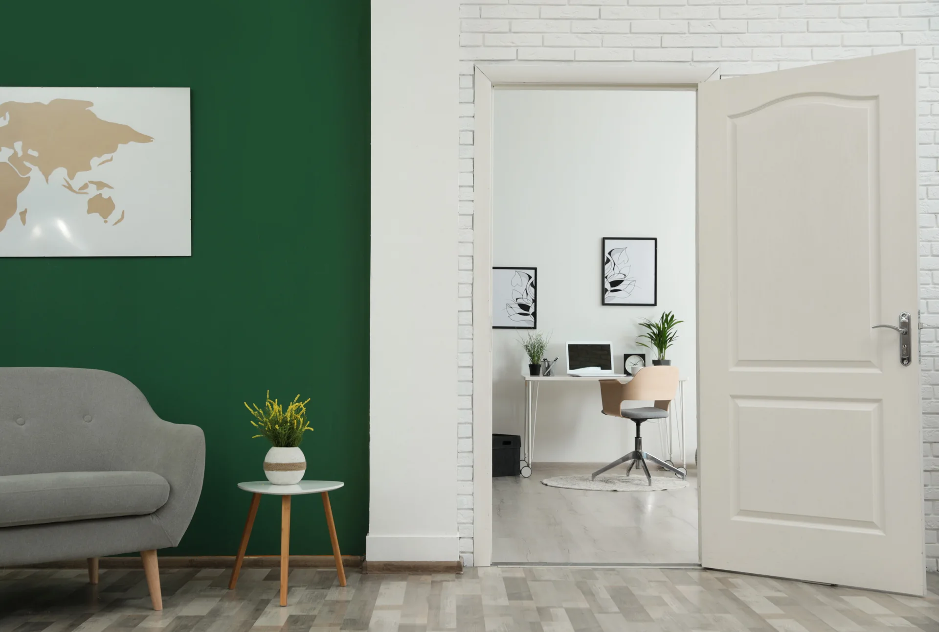 An open doorway framed by a white brick wall leads from a room with a green accent wall to a bright home office with a minimalist desk.