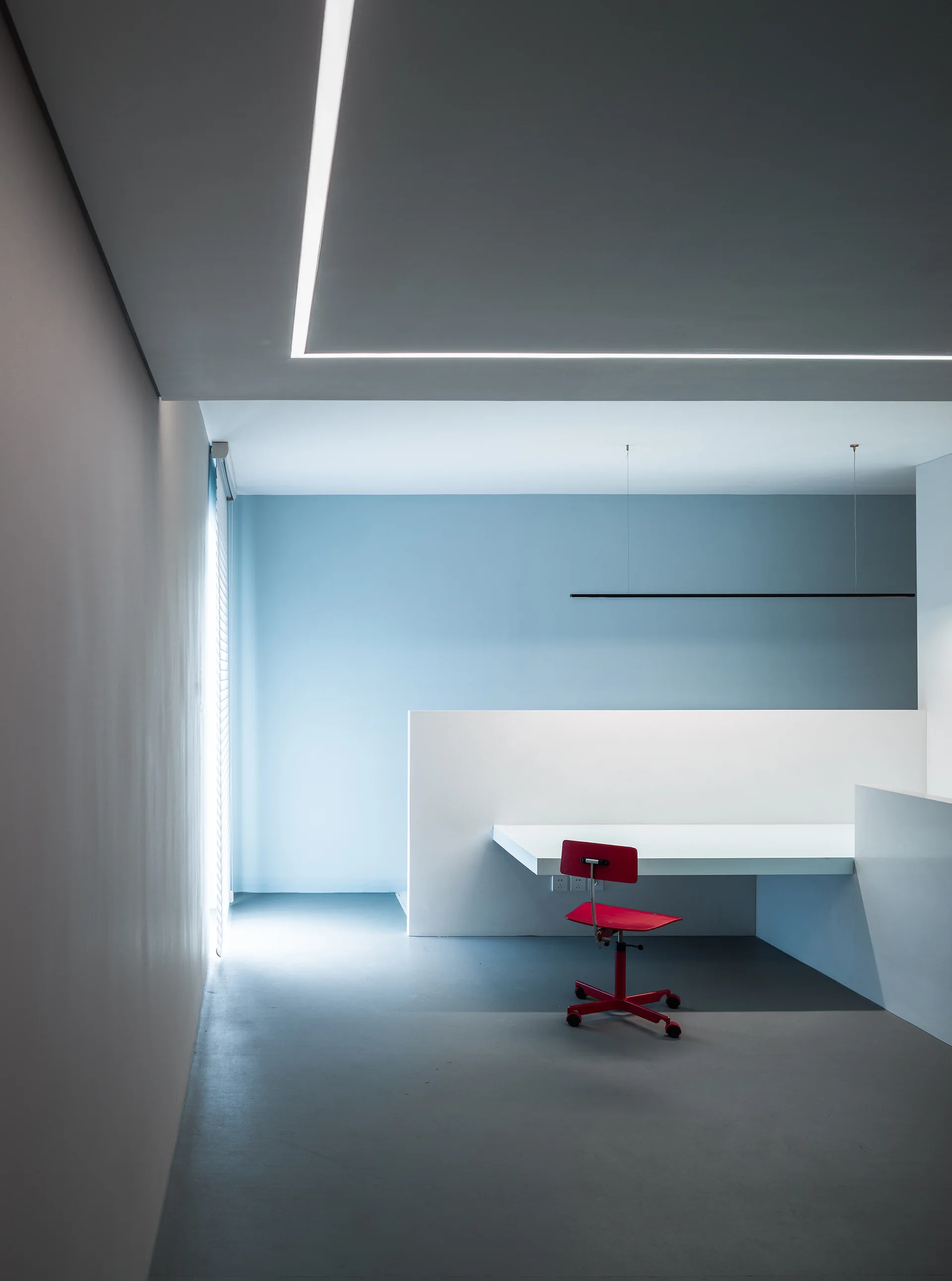 A minimalist home office featuring a white built-in desk, a red wheeled task chair, pale blue walls, and a recessed linear ceiling light strip.