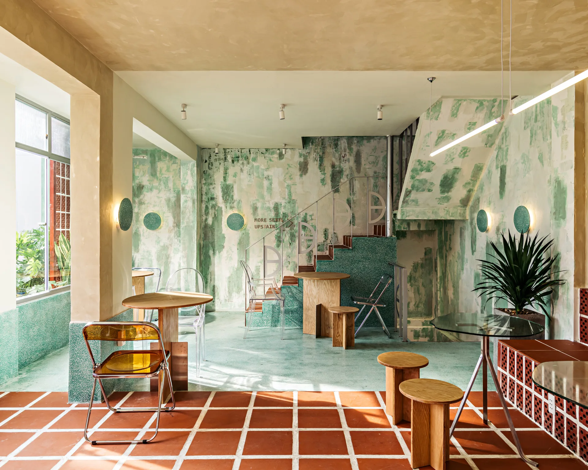 A modern cafe interior featuring washed green walls, a terracotta tiled floor, wood tables, and a geometric metal stair railing.