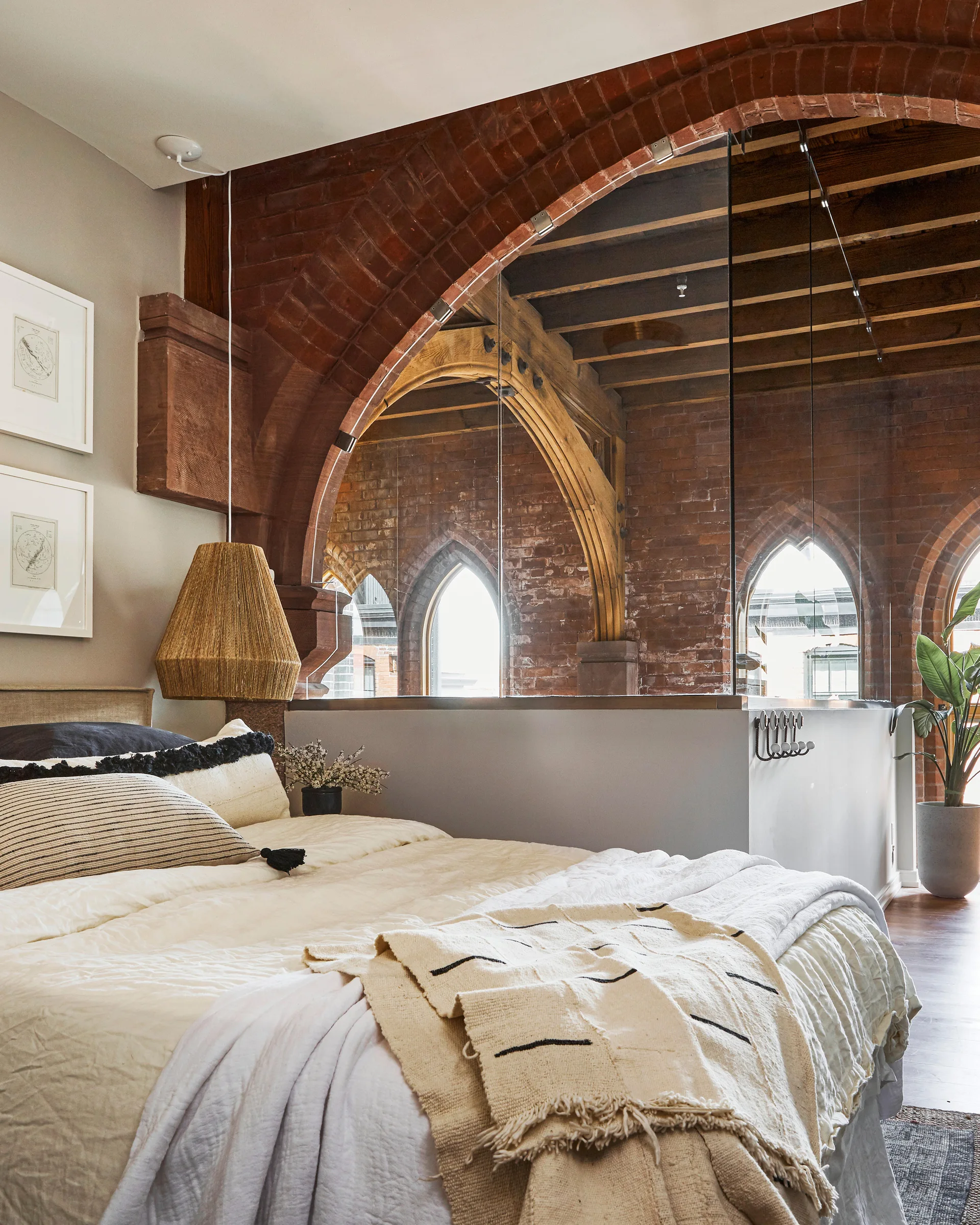 A bedroom inside an industrial loft features a bed with neutral linens, a large red brick archway, and a clear glass partition wall.