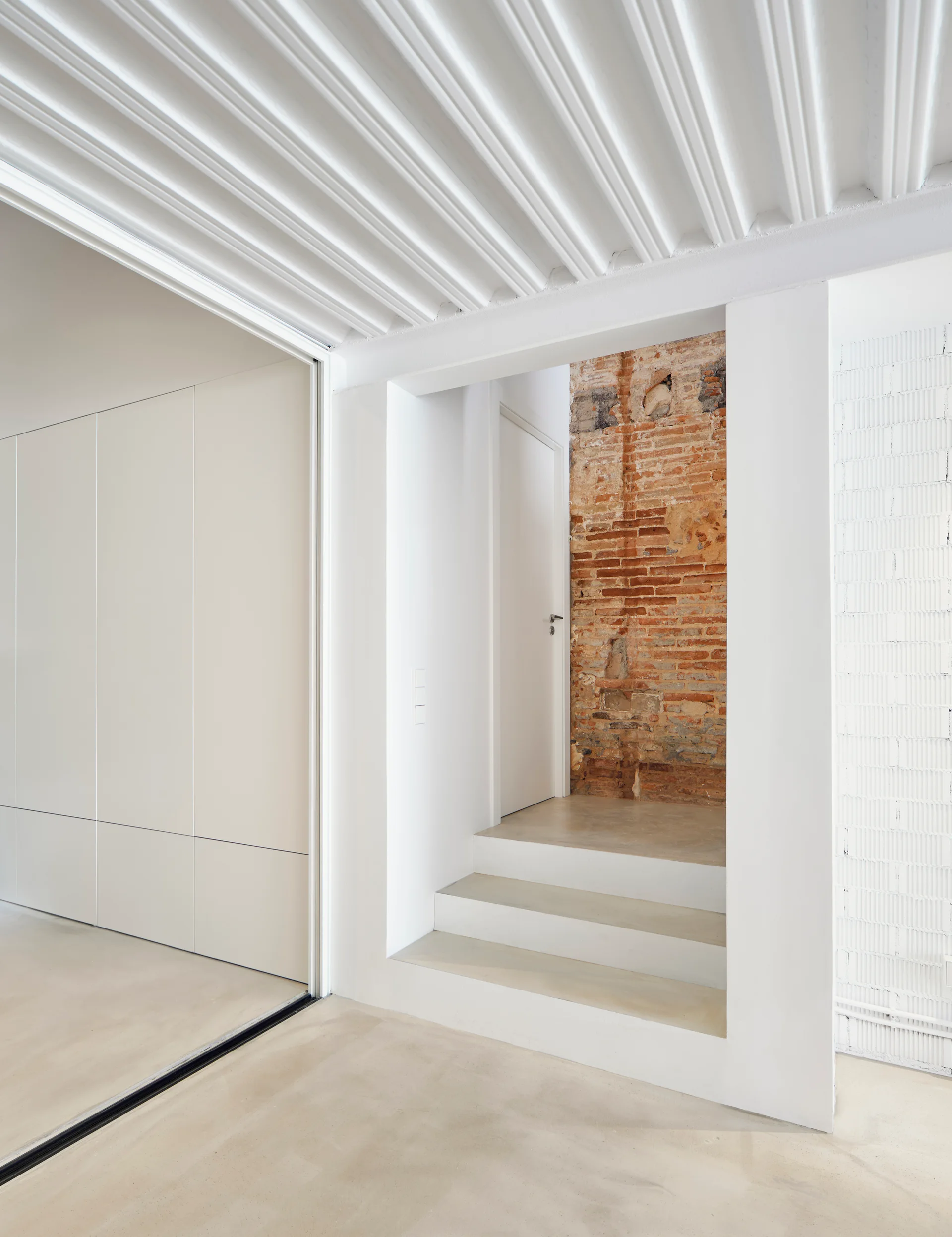 A minimalist hallway features smooth concrete stairs leading to a doorway, set against a backdrop of an exposed brick wall and a white corrugated ceiling.