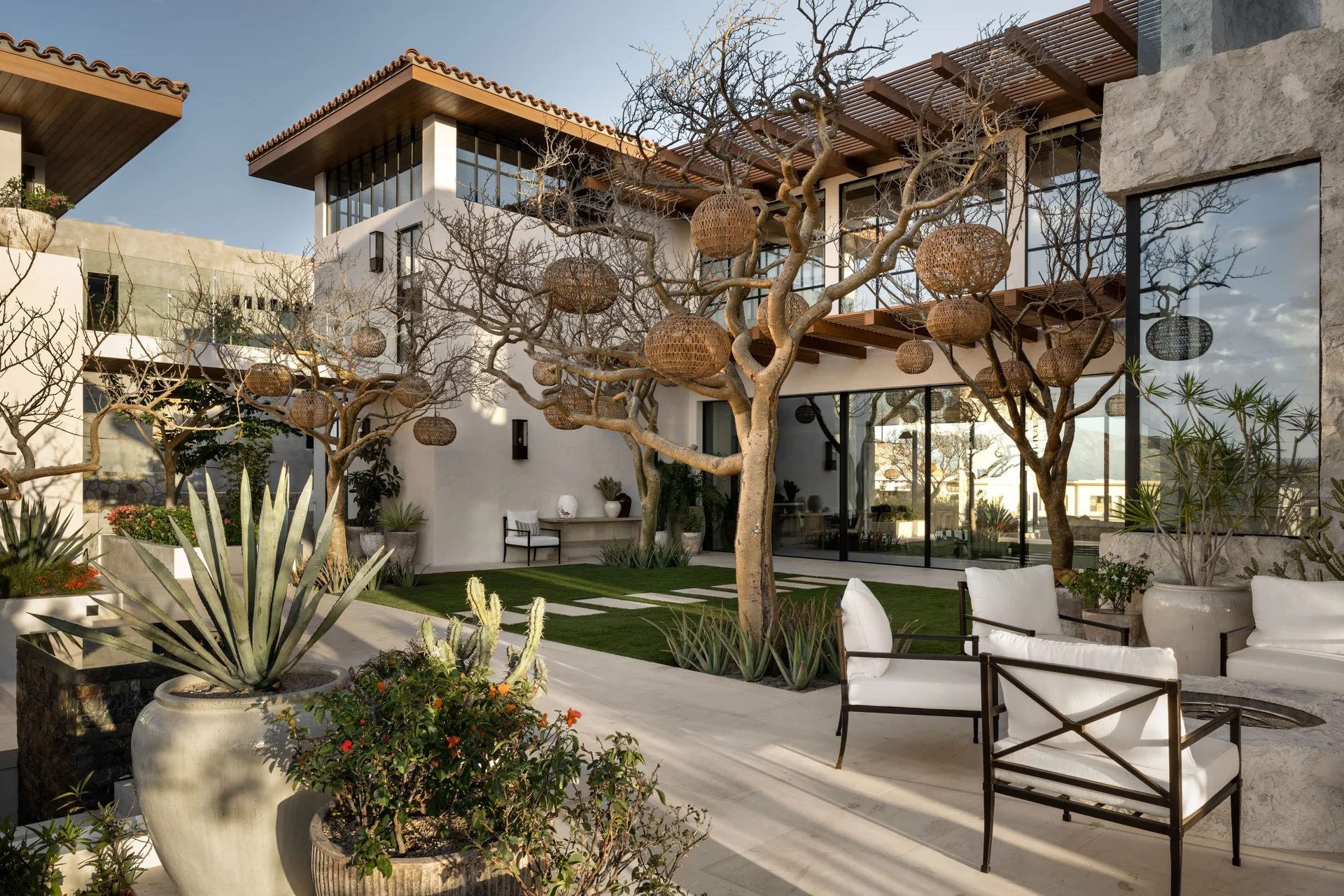 A modern courtyard patio features white lounge chairs around a stone fire pit with woven spherical lights hanging from leafless trees.