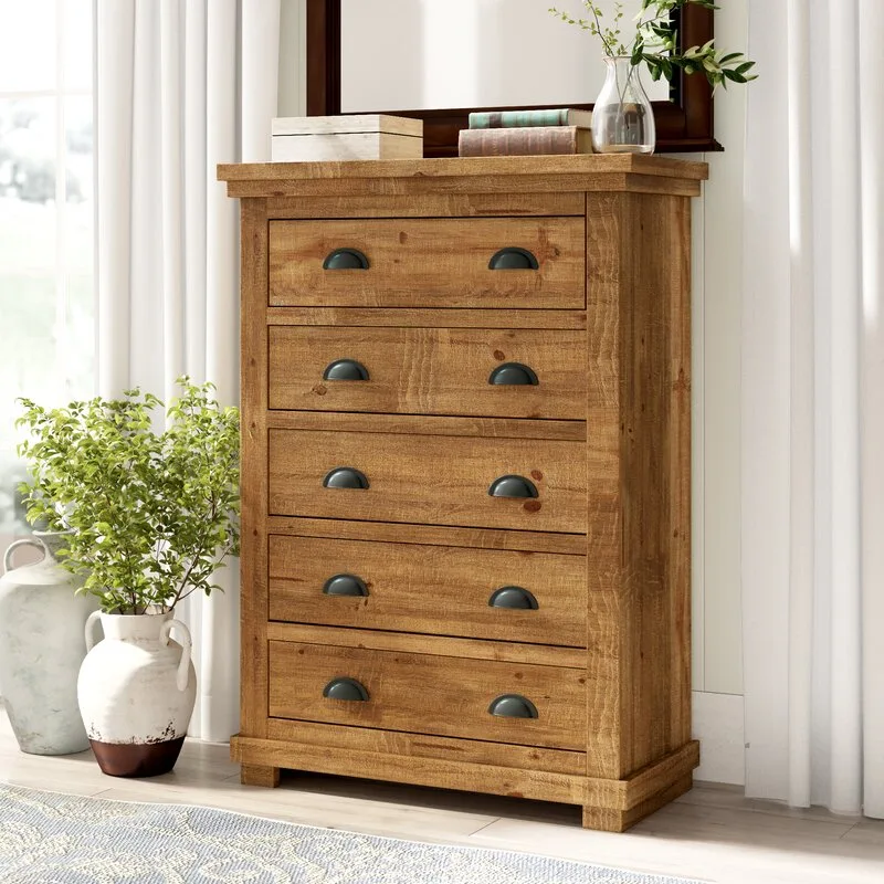 A five-drawer rustic wood dresser with dark metal cup pulls stands in a bright room beside white curtains and a ceramic floor vase.