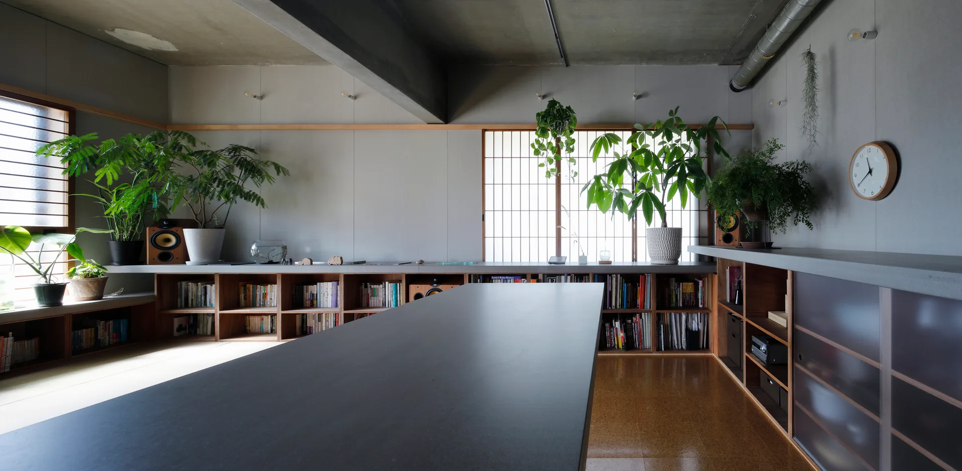 A minimalist room featuring a large dark table, low wood bookshelves built along grey walls, and glowing paper shoji screens under a concrete ceiling.