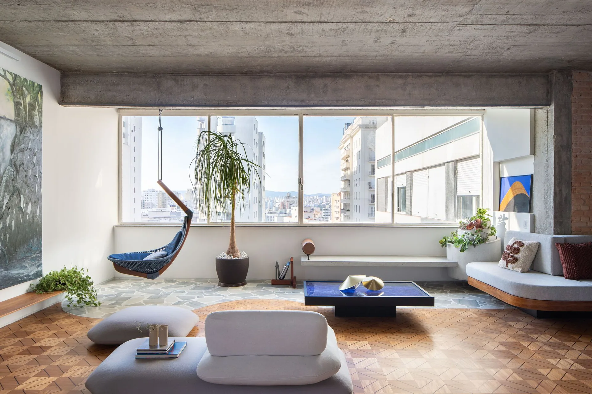 A modern living room featuring mixed wood and stone floors, an exposed concrete ceiling, low-profile furniture, and a window overlooking the city.
