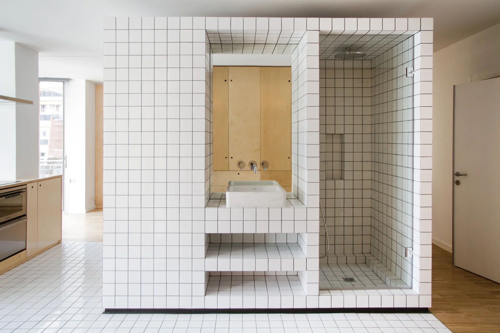 A freestanding bathroom partition block covered in white square grid tiles features an integrated vessel sink and a glass-enclosed walk-in shower.