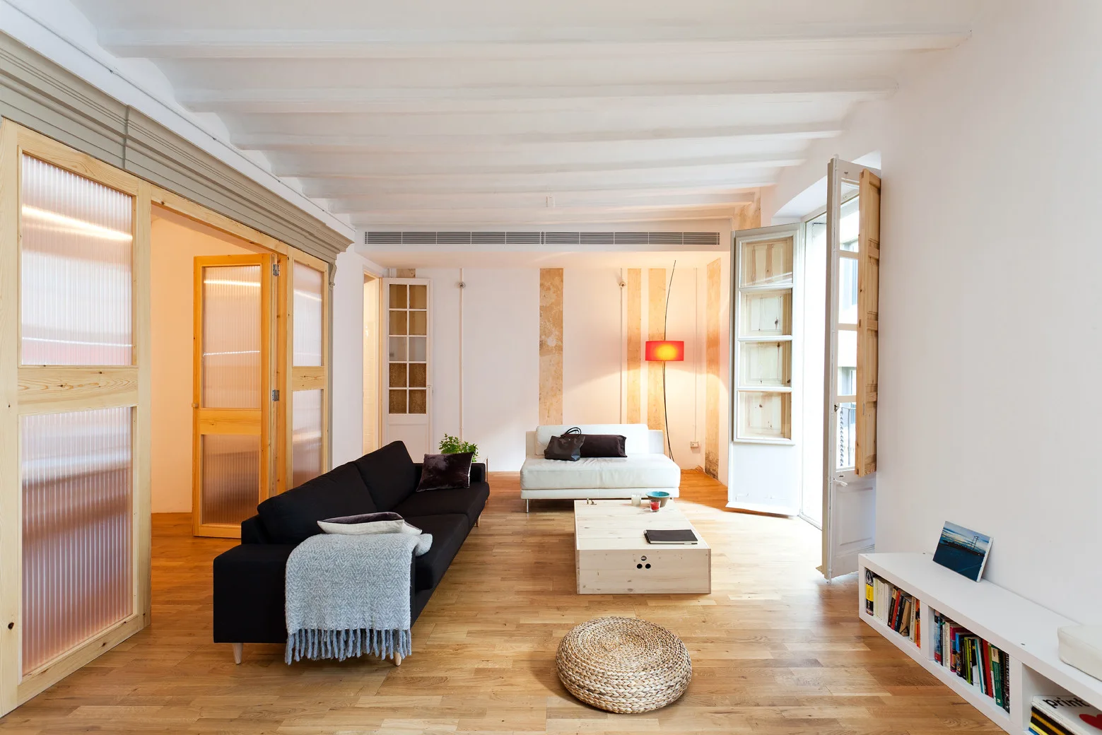 A bright living room features a black sofa, a light wood coffee table, fluted glass sliding doors, and white painted ceiling beams.