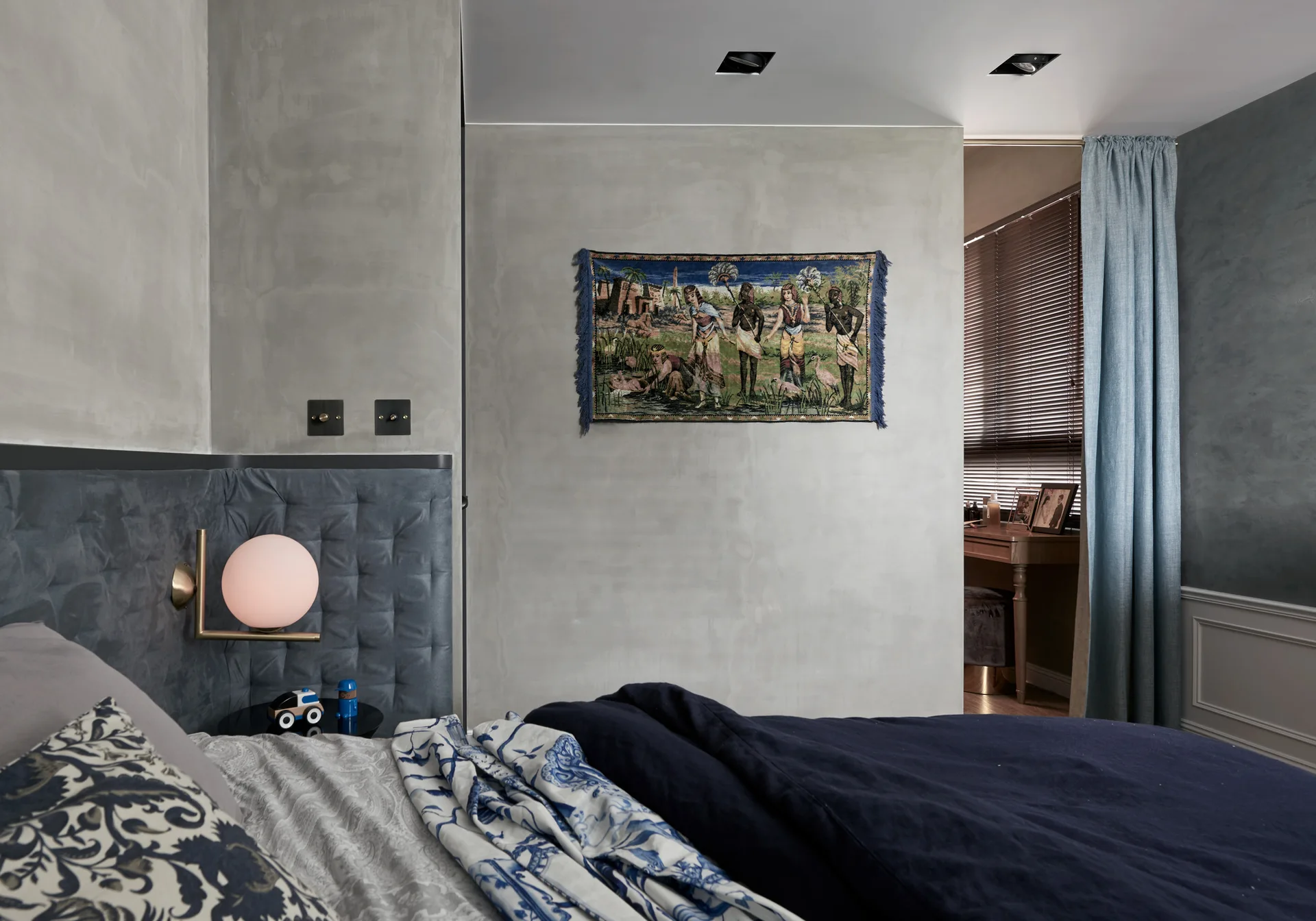 A contemporary bedroom features gray plaster walls, a blue tufted headboard panel with a globe wall sconce, and a small decorative wall tapestry.