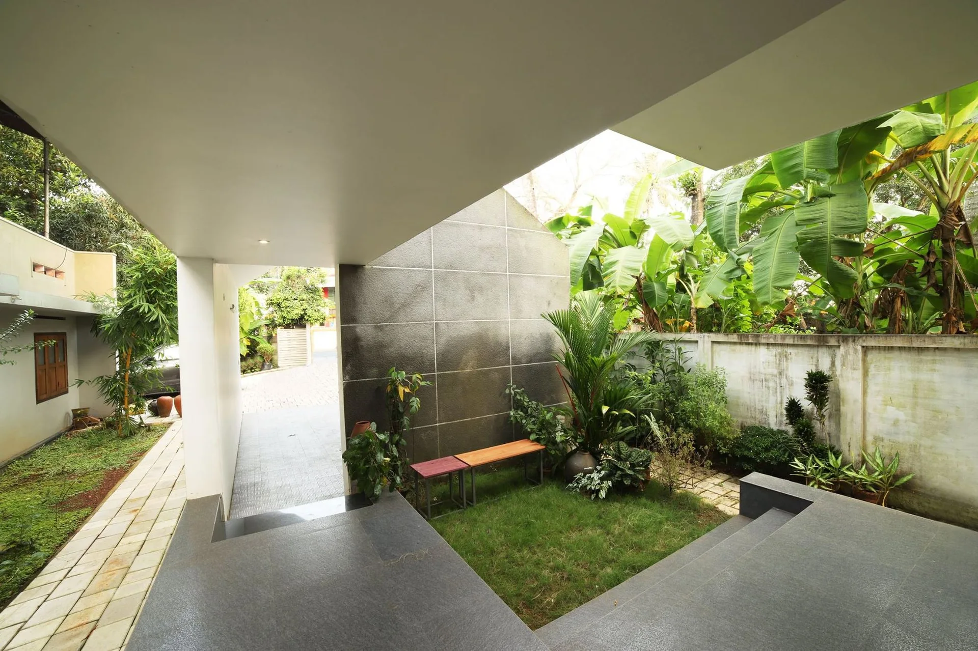 A modern semi-enclosed courtyard featuring a small grass lawn, a tall dark grey tiled wall, a wooden bench, and tropical foliage.