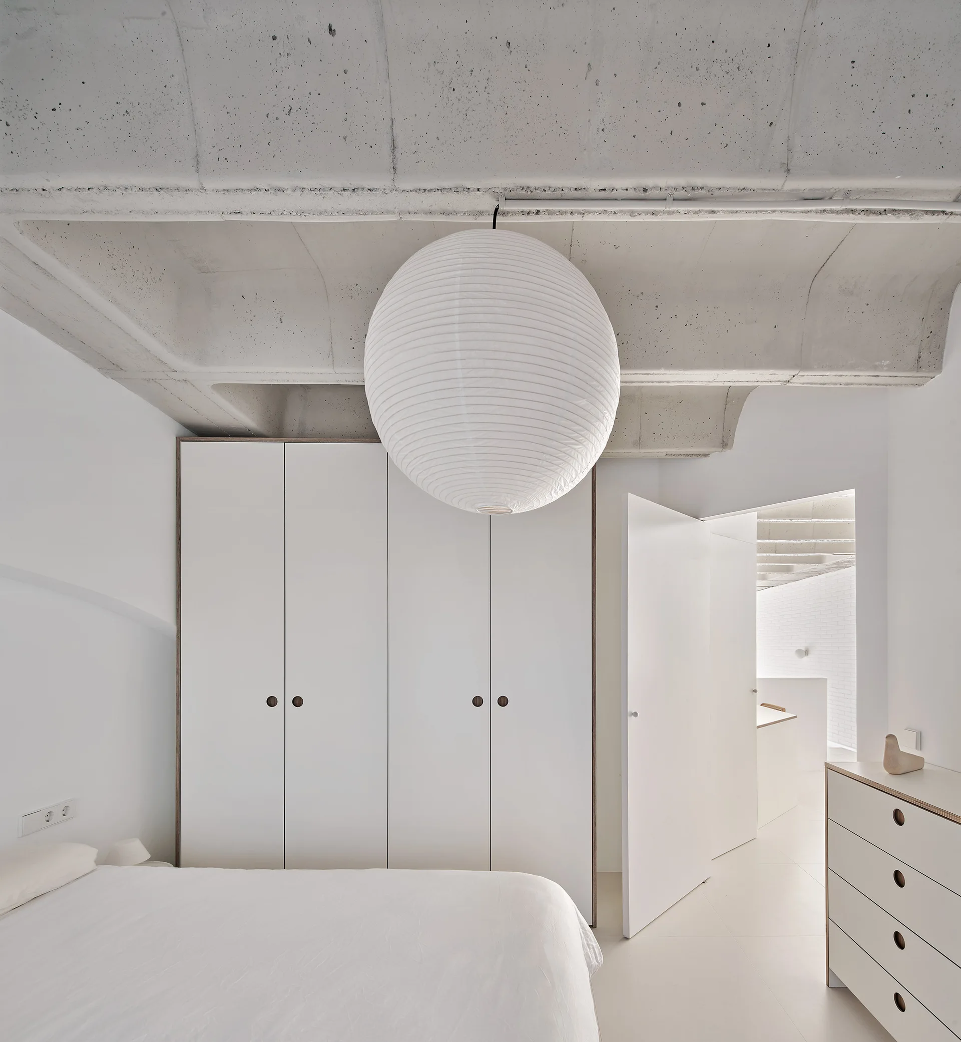 A minimalist bedroom with a large round paper pendant light hanging below a raw concrete ceiling, next to white built-in wardrobes.