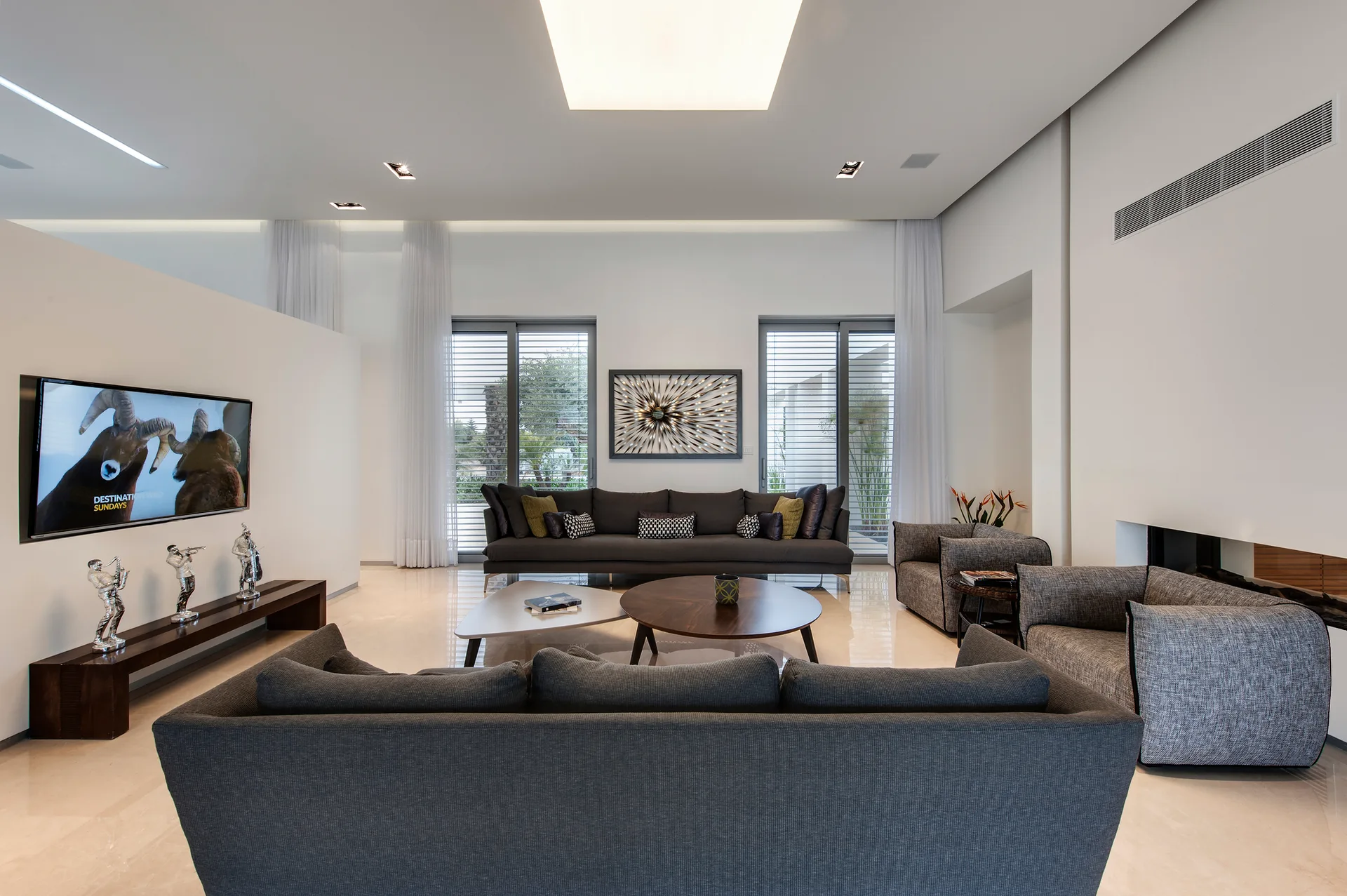 A modern living room features charcoal grey sofas arranged on a polished beige floor around a round wood coffee table and a triangular metal table.