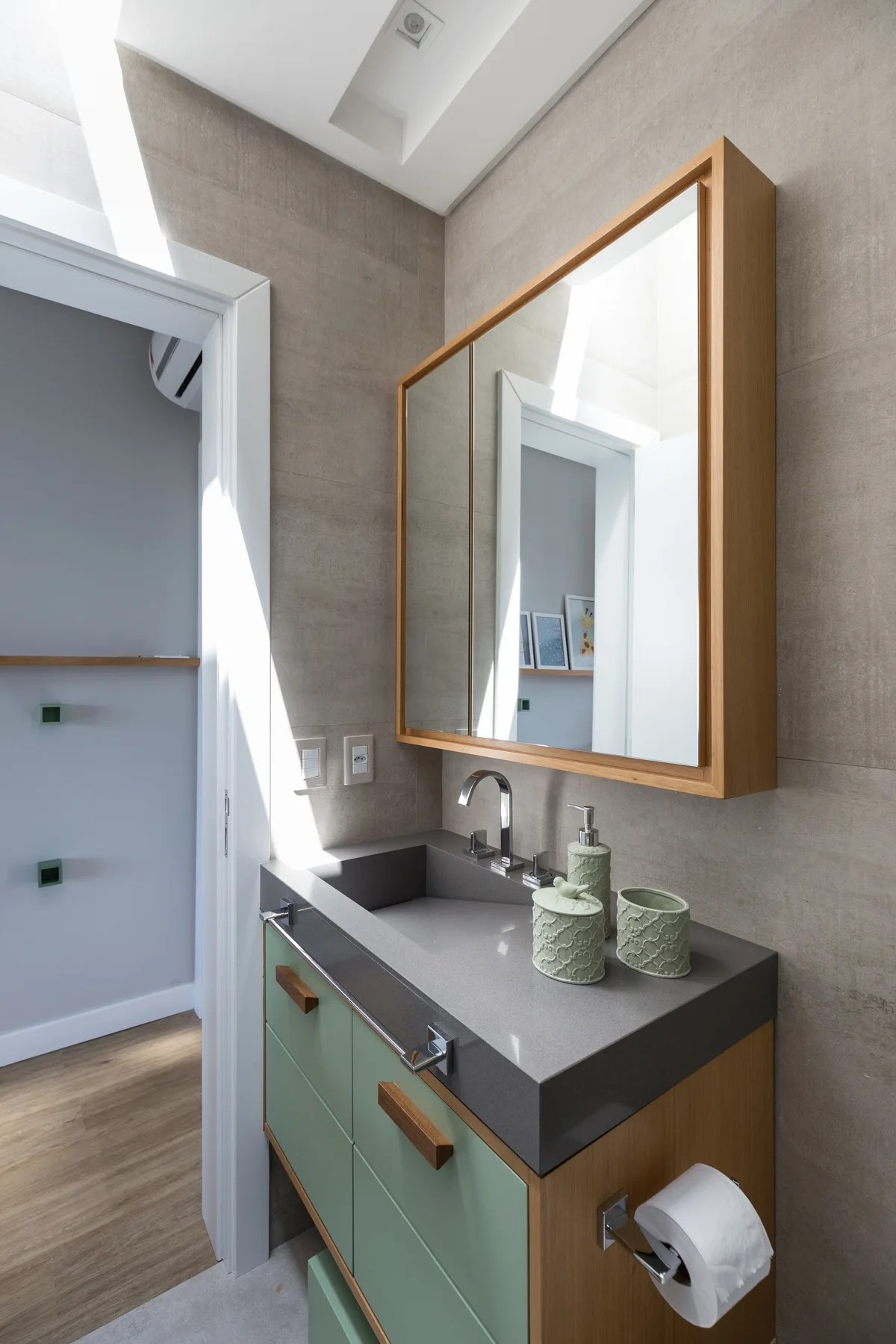 A bathroom vanity features sage green cabinets, a grey countertop with a chrome faucet, and a wood-framed mirrored cabinet on a grey tiled wall.