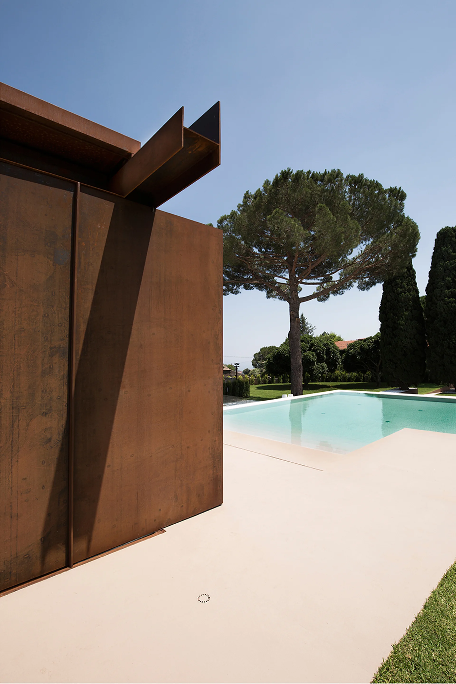 A textured rust-brown steel wall with a projecting beam stands next to a smooth concrete pool deck and a turquoise swimming pool lined with trees.