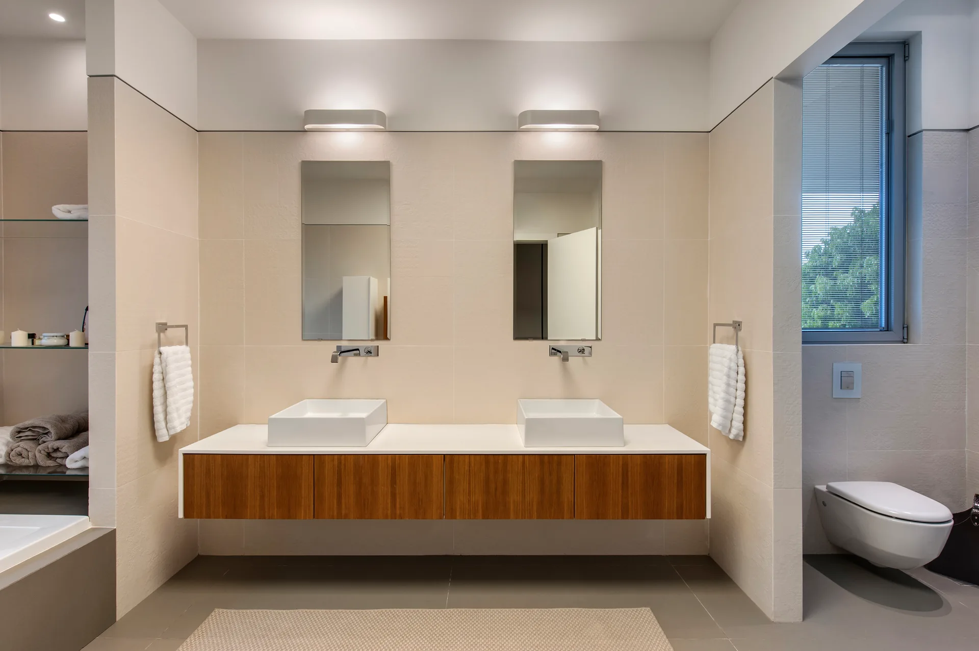 A symmetrical modern bathroom features a floating wood double vanity with square vessel sinks, rectangular mirrors, and beige tiled walls.
