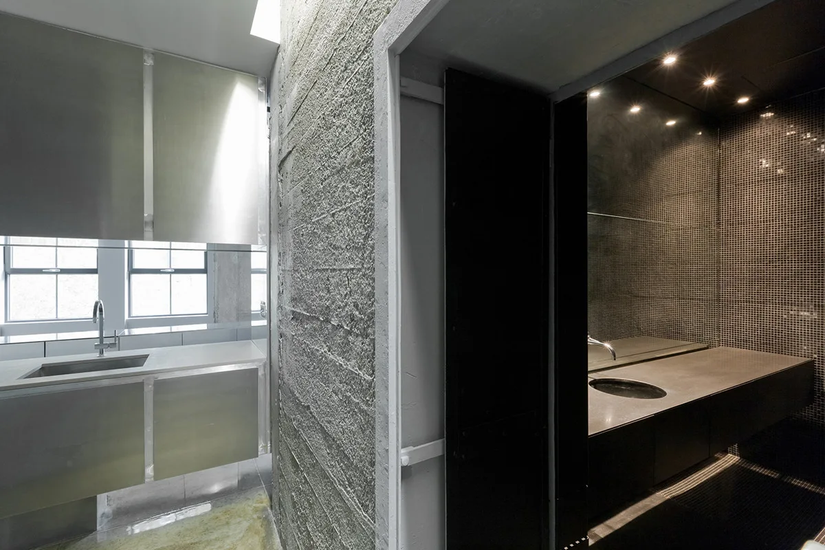 An industrial bathroom featuring a textured concrete wall, a stainless steel vanity, and a dark mosaic tiled sink area with recessed lighting.