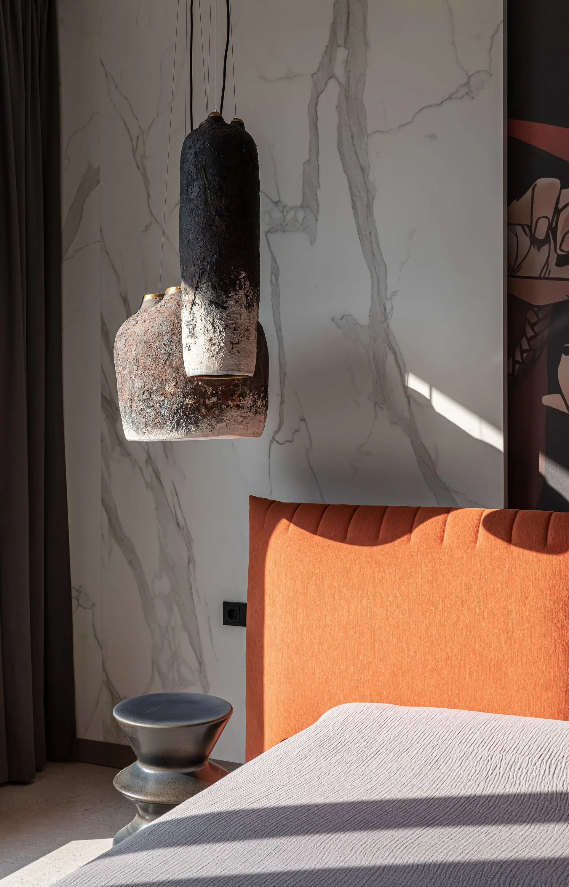 A close-up of a bed with an orange upholstered headboard against a white marbled wall panel next to rough textured pendant lights.