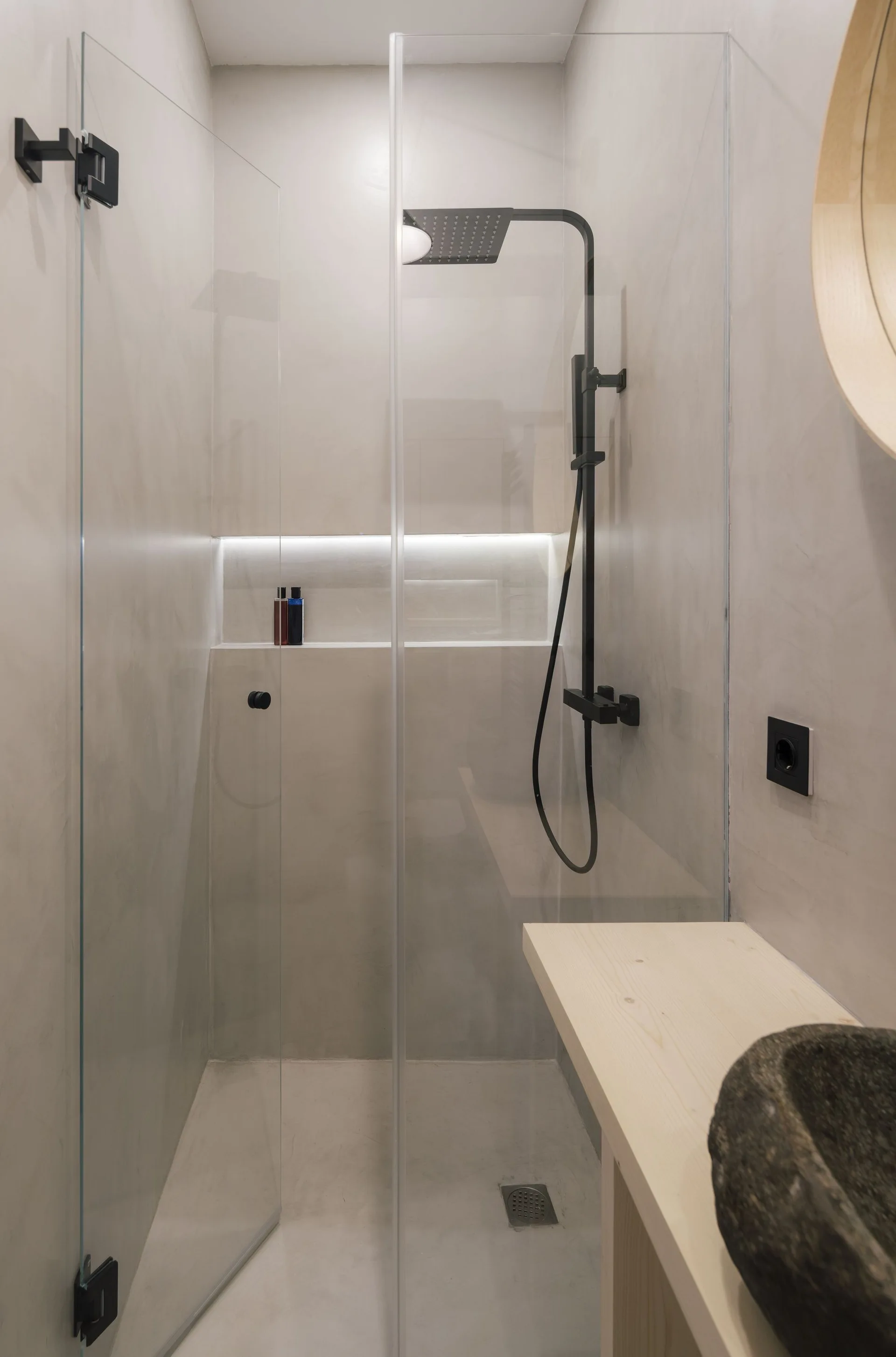 A modern shower with grey plaster walls, a matte black shower head, a glass screen, and a recessed backlit wall niche.