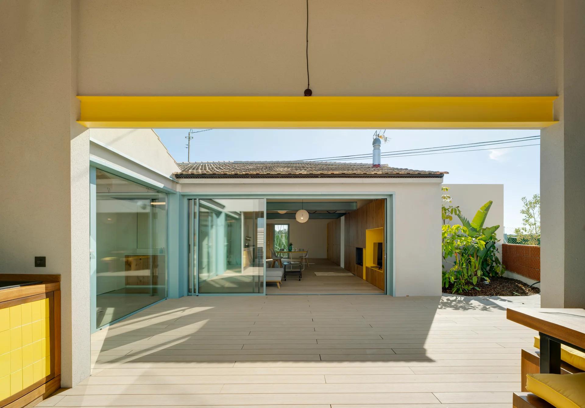 An outdoor plank deck features a bright yellow structural beam framing a view of a wood-paneled interior room through sliding glass doors.