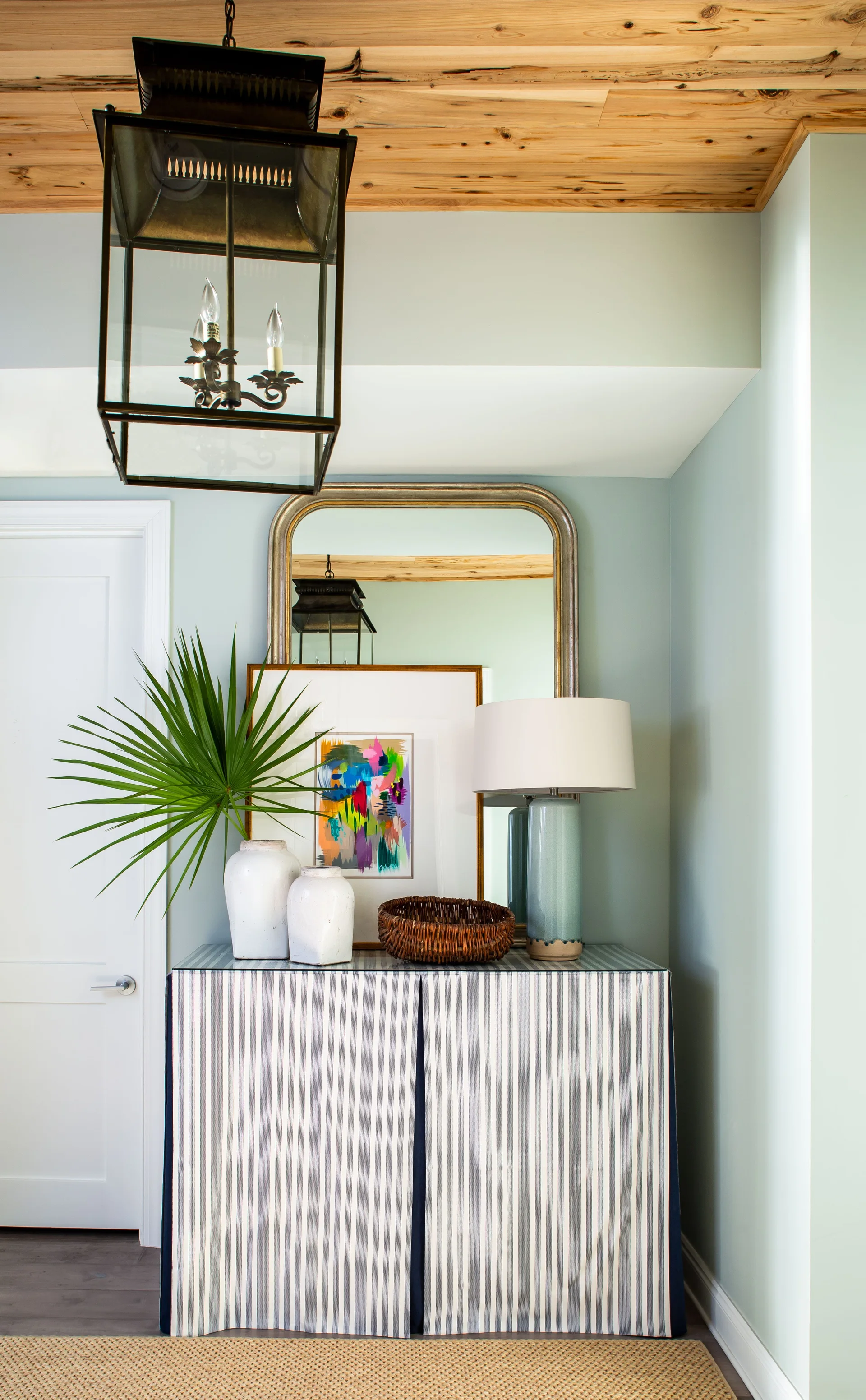 A striped skirted console table stands against a light blue wall beneath a wood plank ceiling and a large black lantern pendant light.