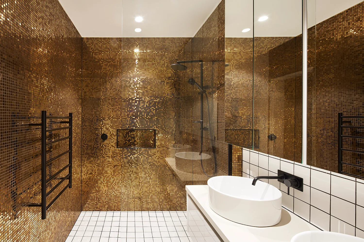 A modern bathroom features metallic gold mosaic tile walls, a white vanity with an oval vessel sink, black plumbing fixtures, and a glass-enclosed shower.