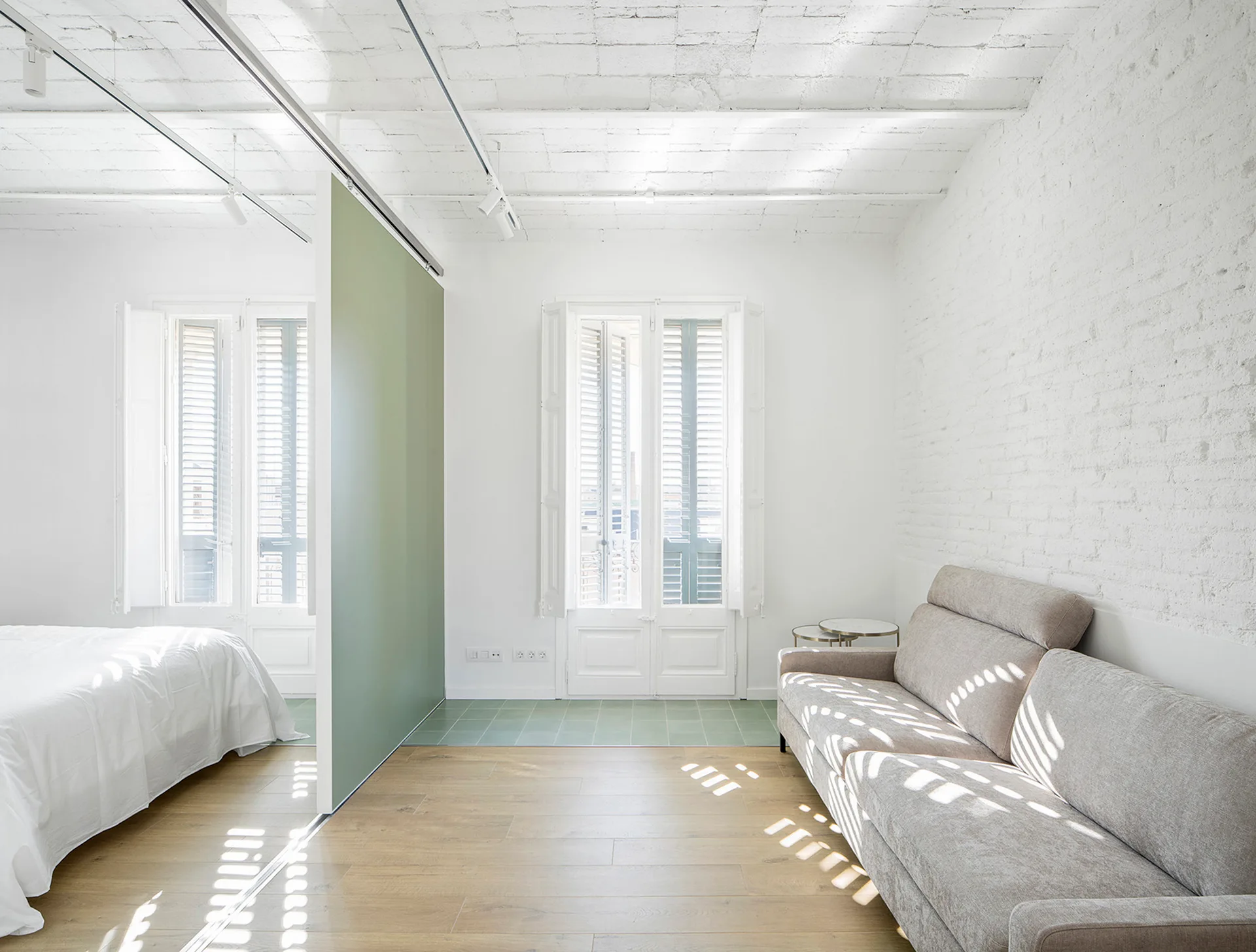 A minimalist studio apartment with a light gray sofa, white brick walls, and a sage green sliding partition separating the bedroom area.