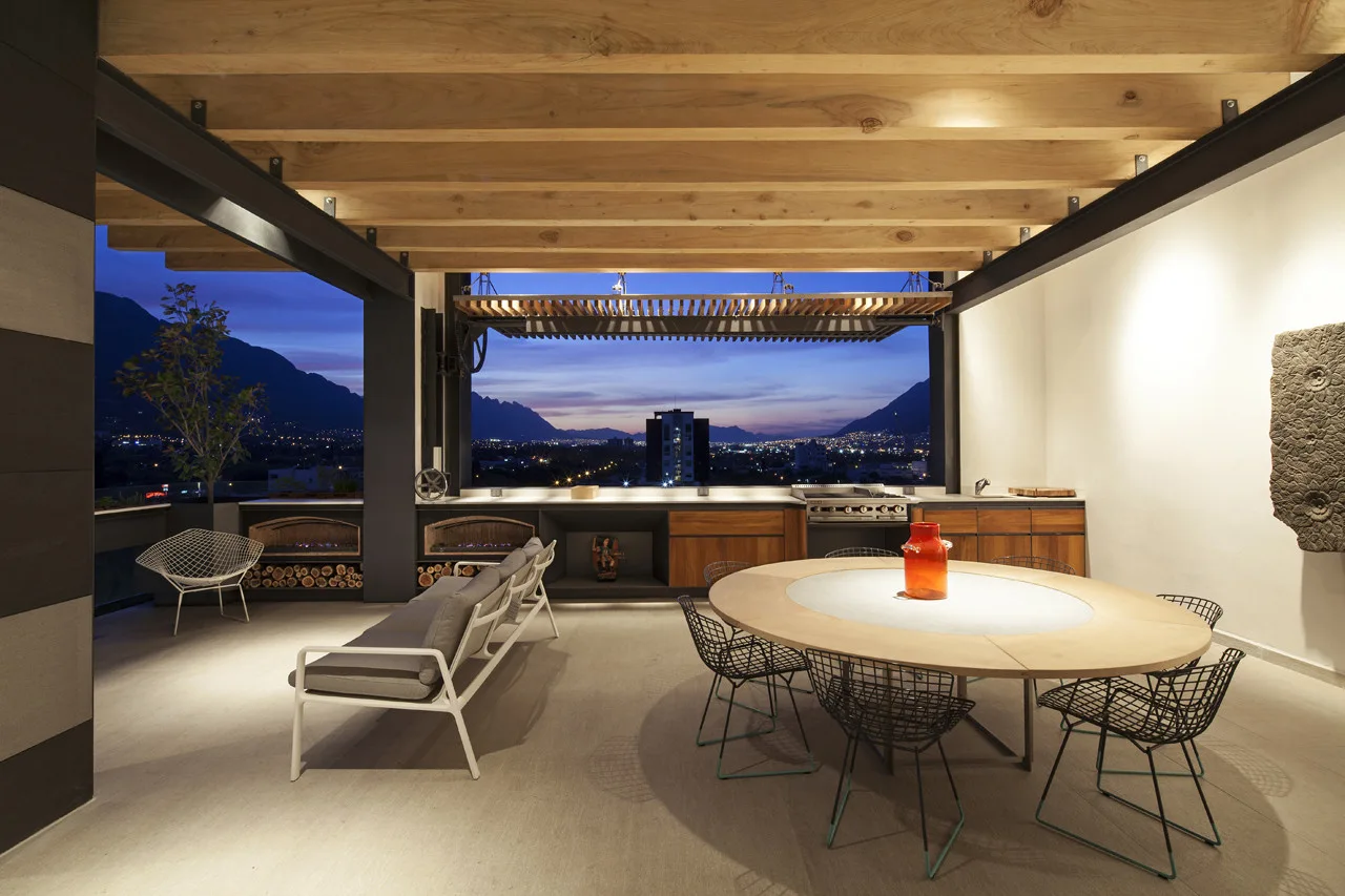 An open-air covered terrace with exposed wood ceiling beams, a round dining table, a grey sofa, and a built-in kitchen overlooking a city skyline.
