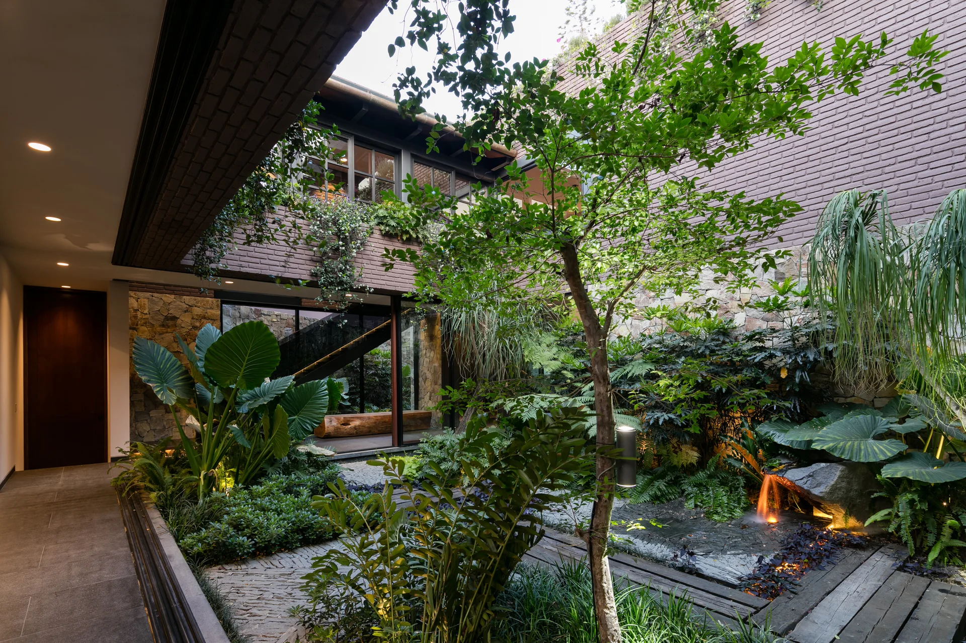 A landscaped interior courtyard garden features lush green tropical plants, brick and stone walls, stone paving, and a small illuminated waterfall.