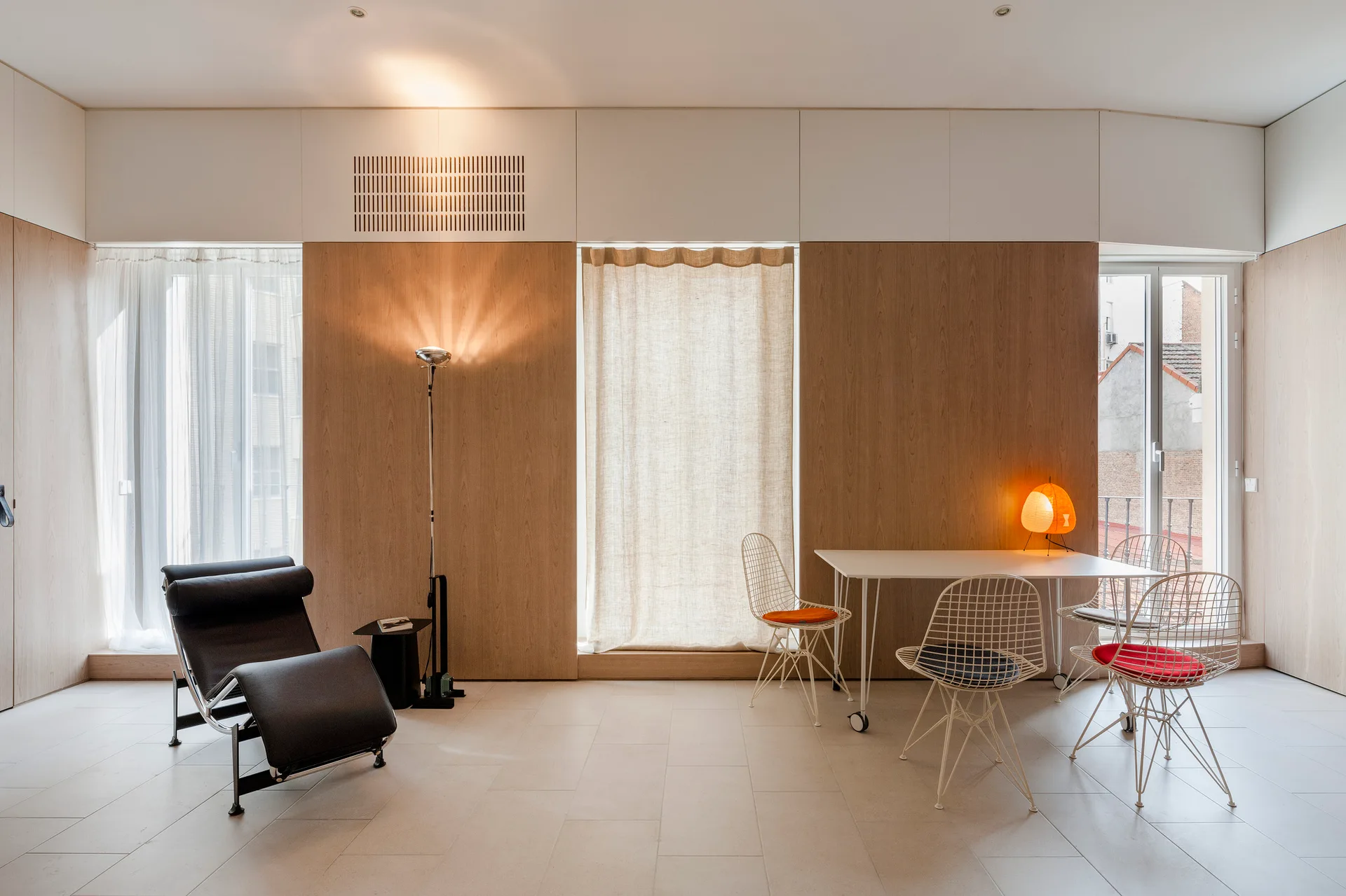 A minimalist living and dining space features large wood wall panels, a black leather lounge chair, and a white dining table with wire frame chairs.