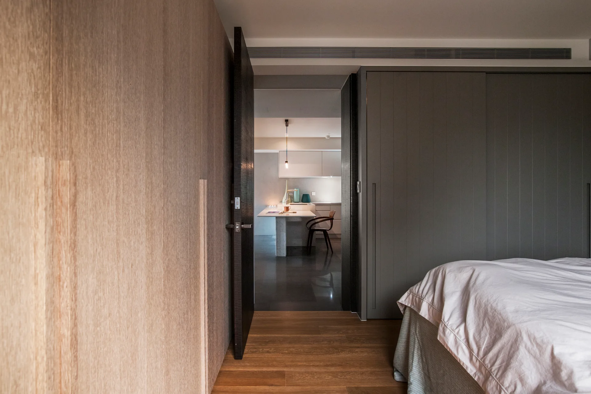 A view from a bedroom with wood-paneled walls and a grey wardrobe looking through an open doorway into a minimalist kitchen with a white island.