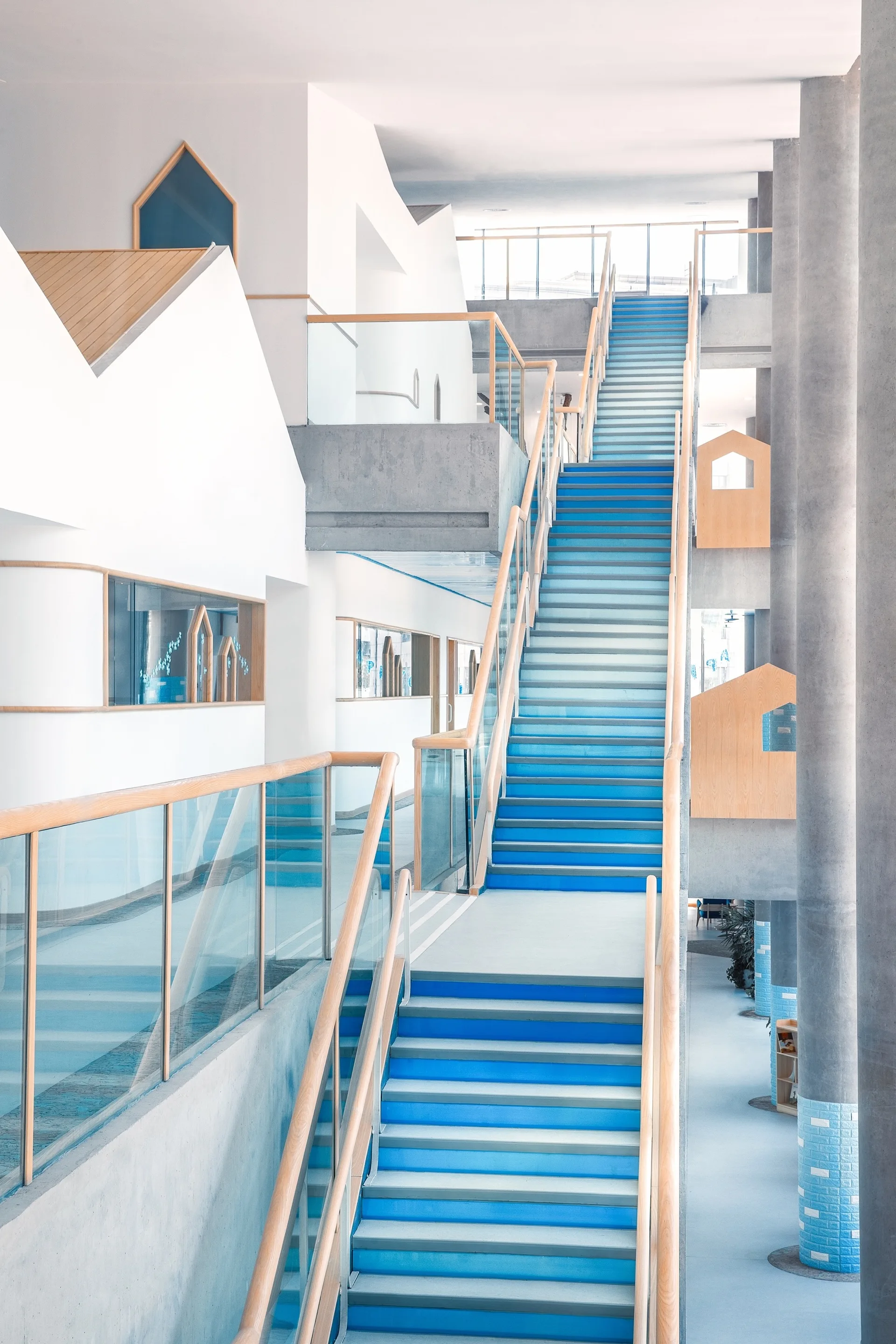 A modern multi-level staircase features bright blue steps, wood and glass handrails, and tall concrete support columns against white walls.
