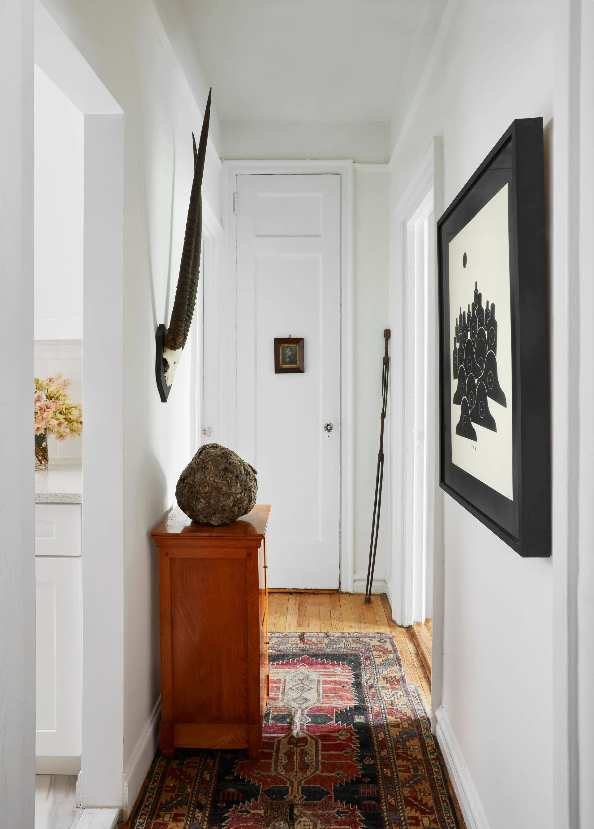 A narrow white hallway featuring a varnished wood console cabinet, a patterned runner rug, and eclectic black framed artwork on the wall.