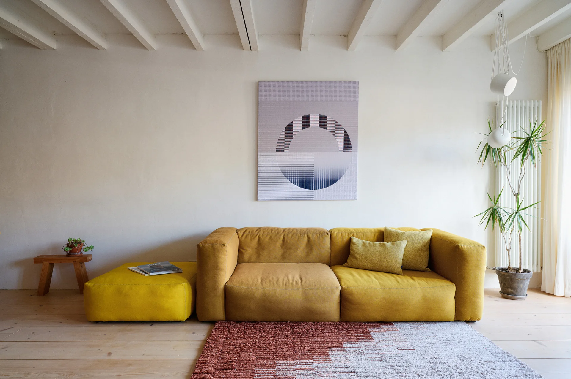 A modern living room features a low yellow sofa and matching ottoman on a textured geometric rug beneath white painted ceiling beams.