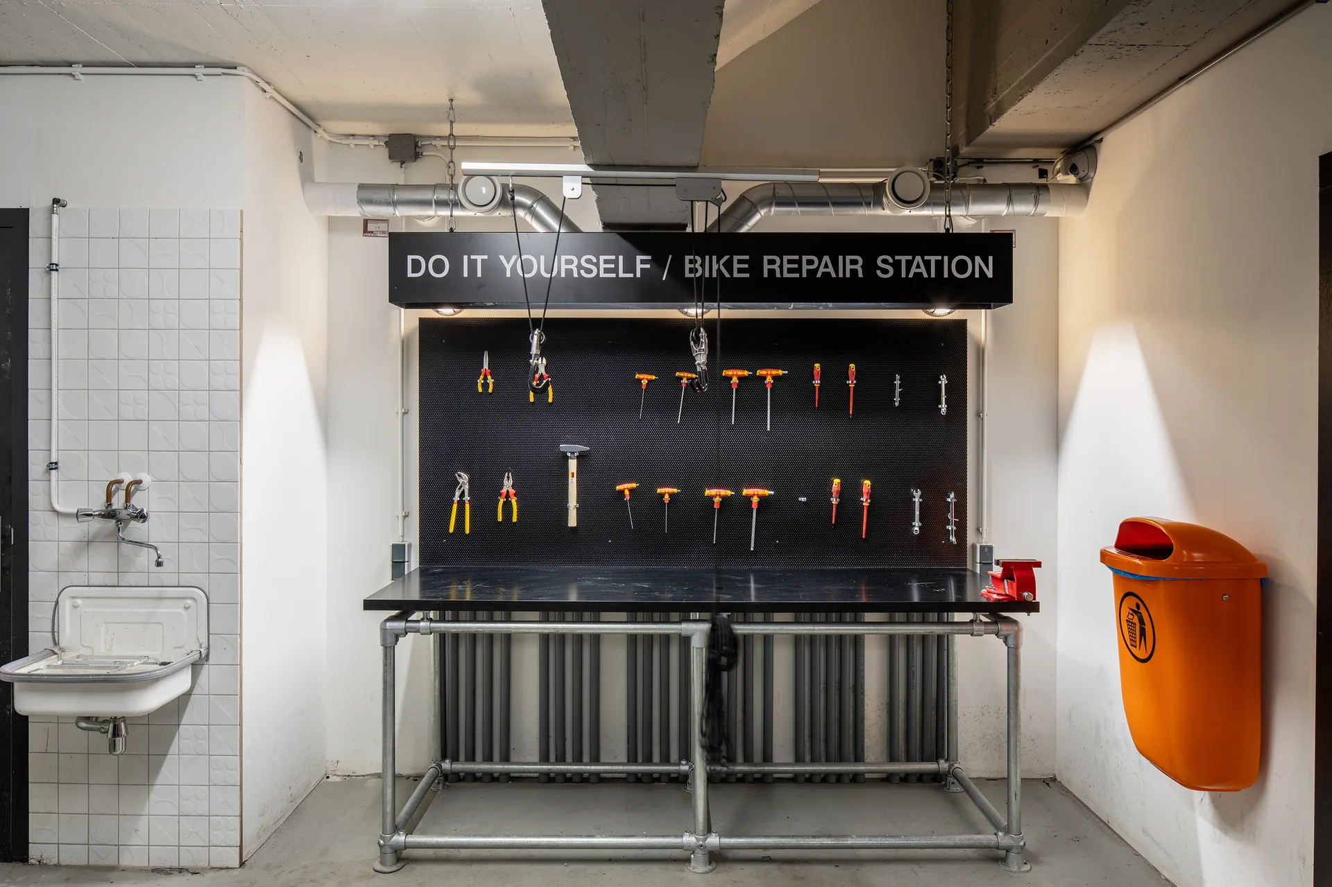 An industrial bike repair station features a black metal pipe workbench, a matching tool-lined pegboard, and a wall-mounted orange bin.