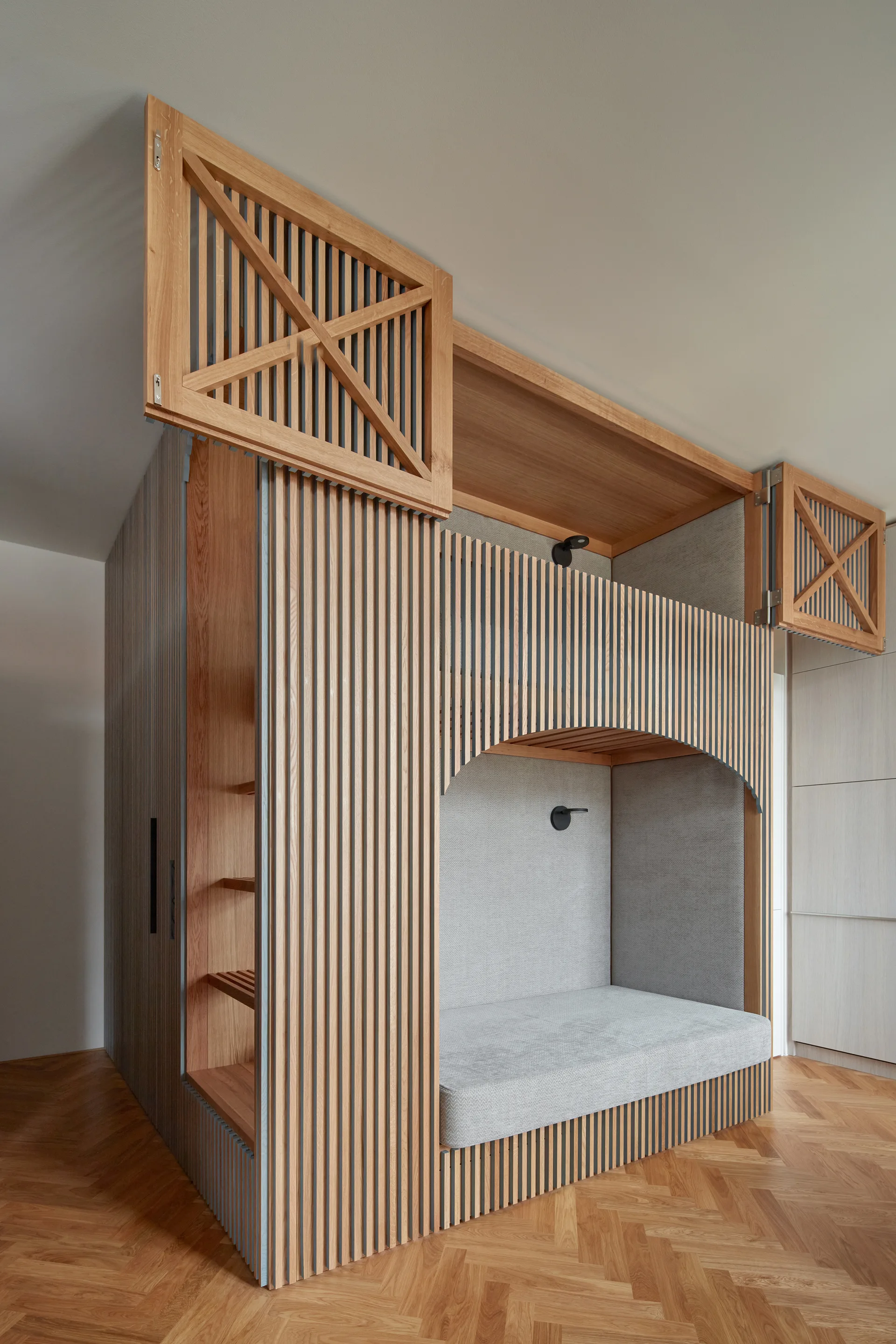 A custom light wood bunk bed structure features vertical slats, an arched sleeping alcove with grey cushioning, and herringbone parquet flooring.