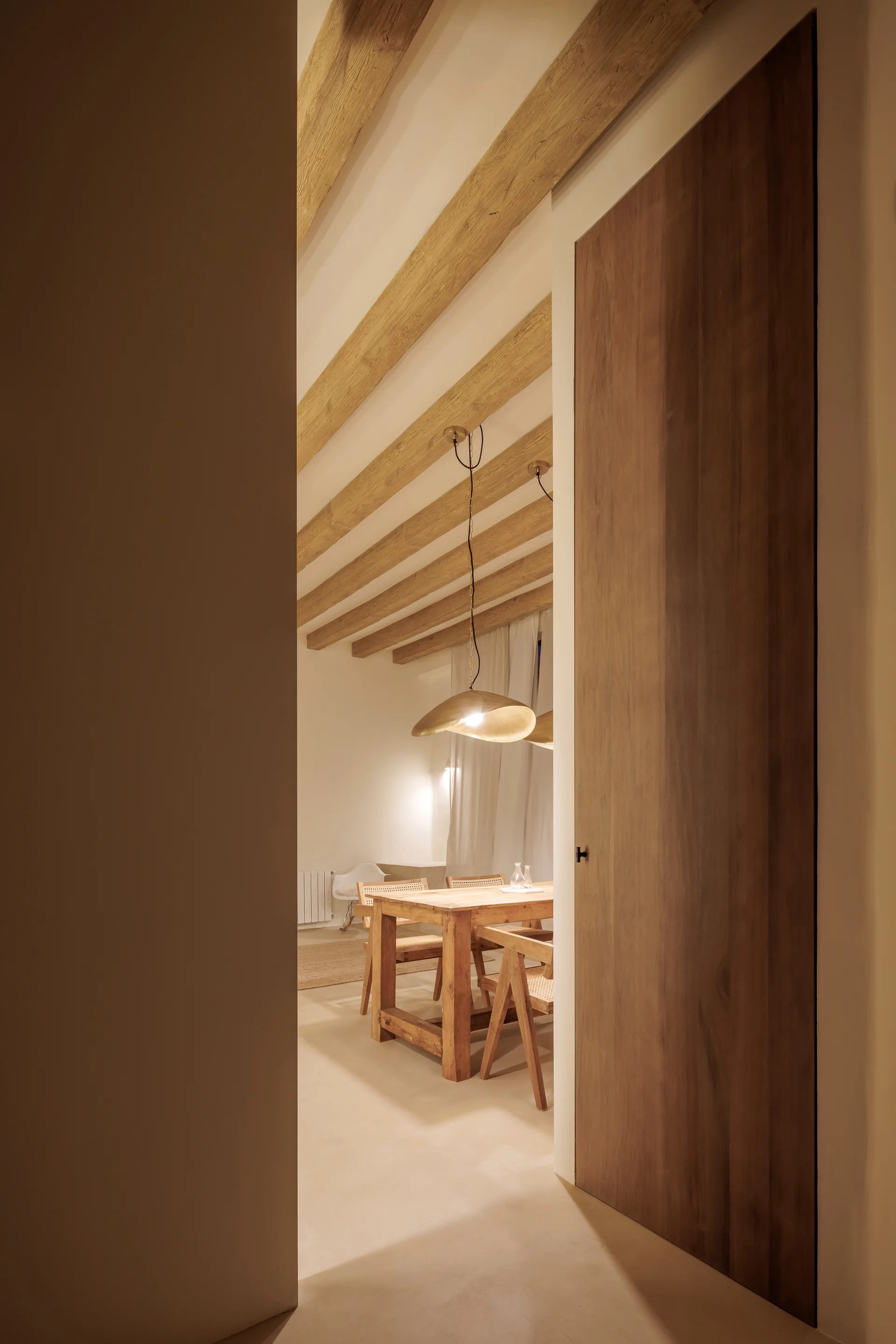 A view through a wooden doorway into a warm dining room featuring a wood table, woven chairs, and rustic exposed ceiling beams.