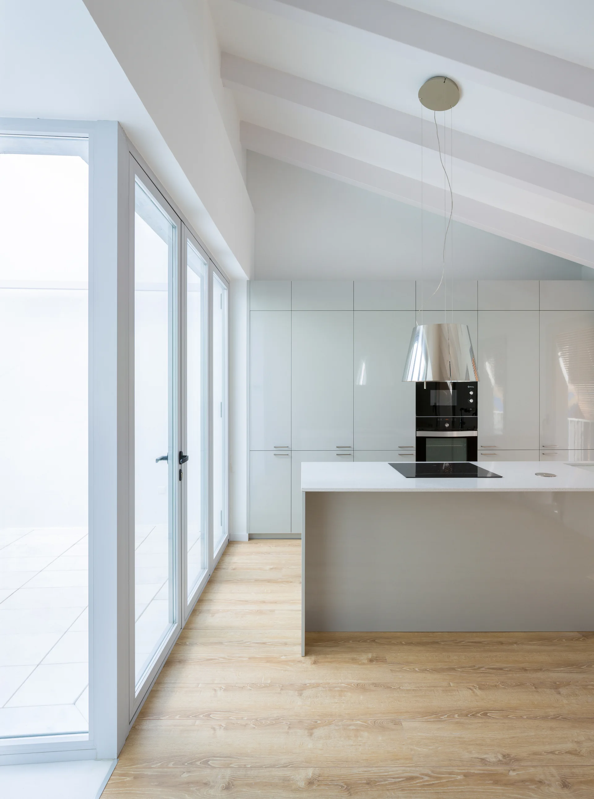 A minimalist kitchen features a light gray island and glossy cabinets under a white vaulted ceiling with exposed beams next to large glass doors.