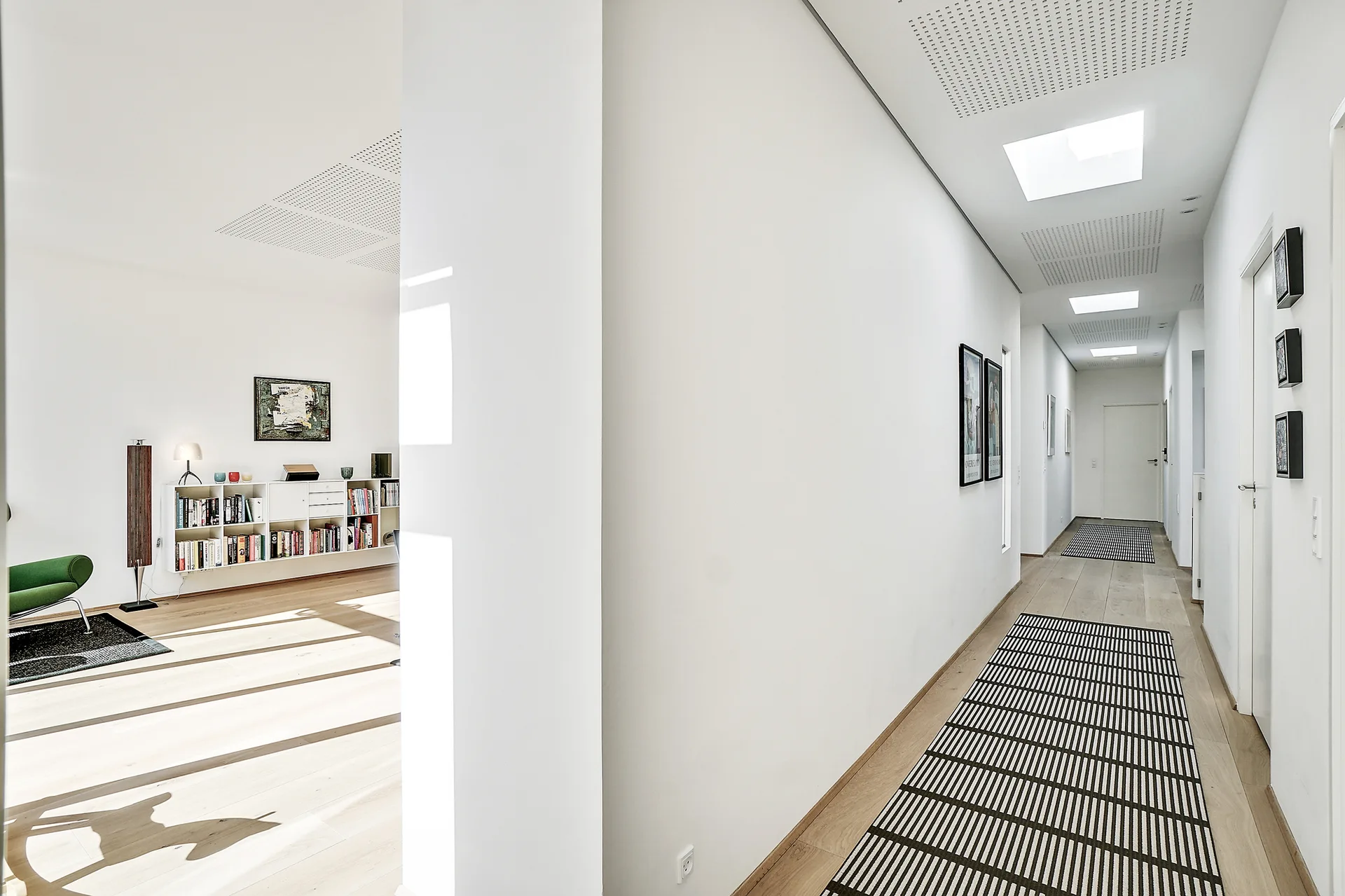 A minimalist white hallway with light wood floors, black and white striped runner rugs, and recessed square ceiling lights leading to a closed door.