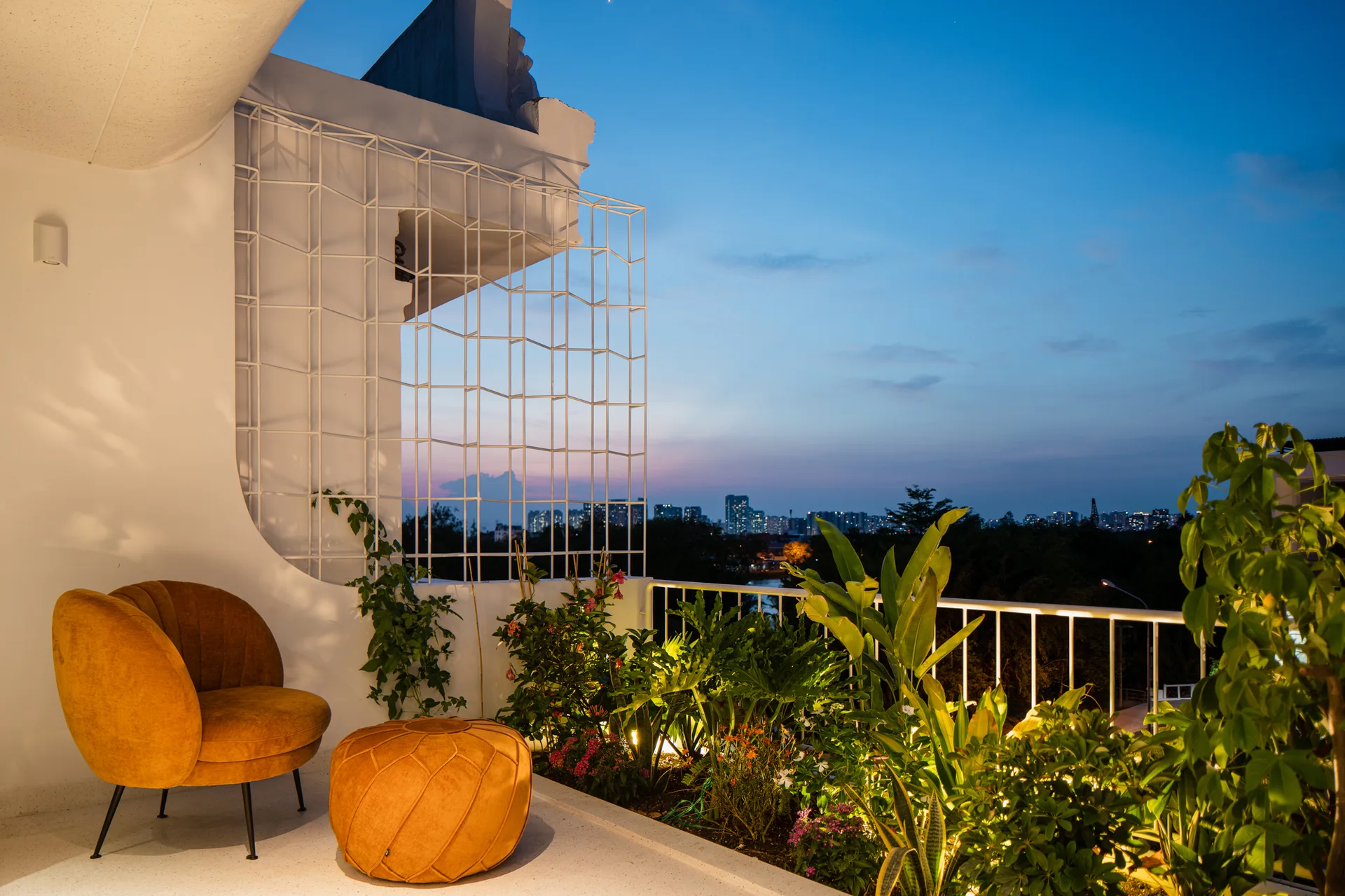 An amber outdoor armchair and matching pouf sit on a white balcony next to a green planter bed and a white metal trellis at twilight.