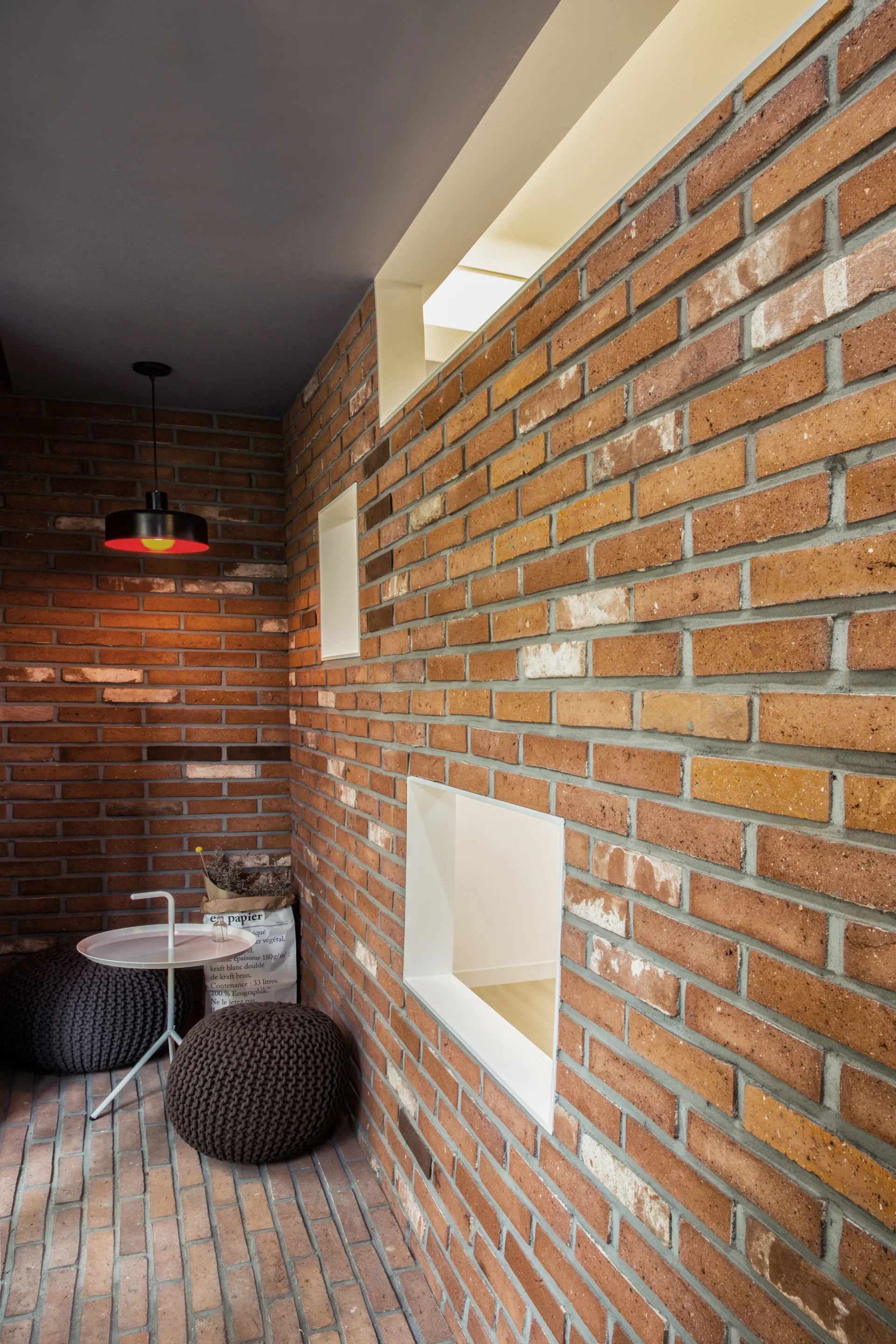 A sitting nook with exposed red brick walls and floors, square recessed white-lined wall openings, knitted poufs, and a small white side table.