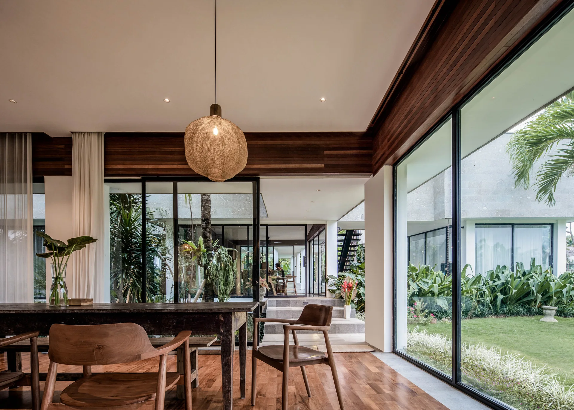 A modern dining room features a dark wood table with matching chairs and a woven pendant light next to expansive glass walls overlooking a green courtyard.