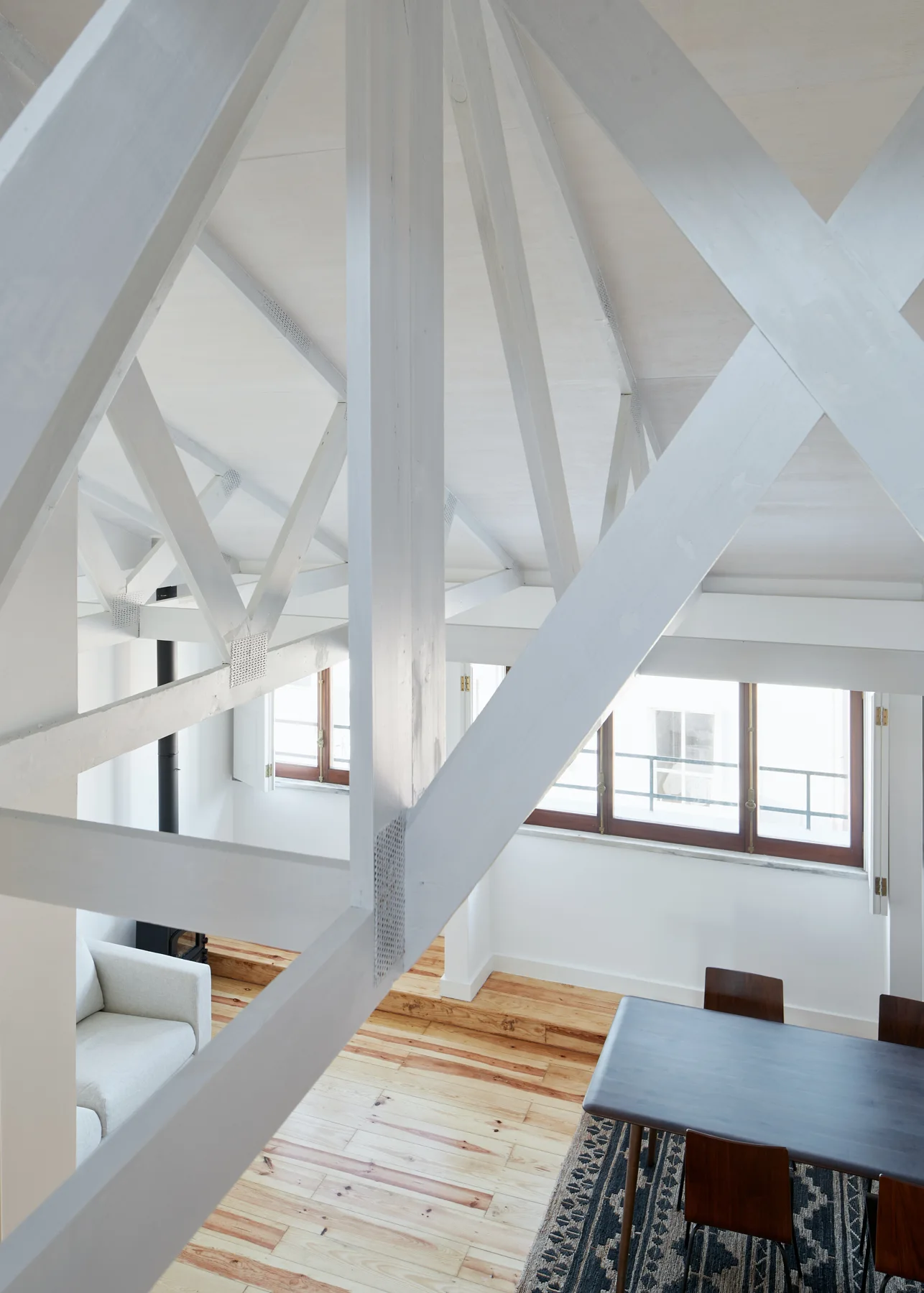 An elevated view of a bright room featuring massive white painted wooden trusses overhead, a dark dining table set on a patterned rug, and pine wood floors.