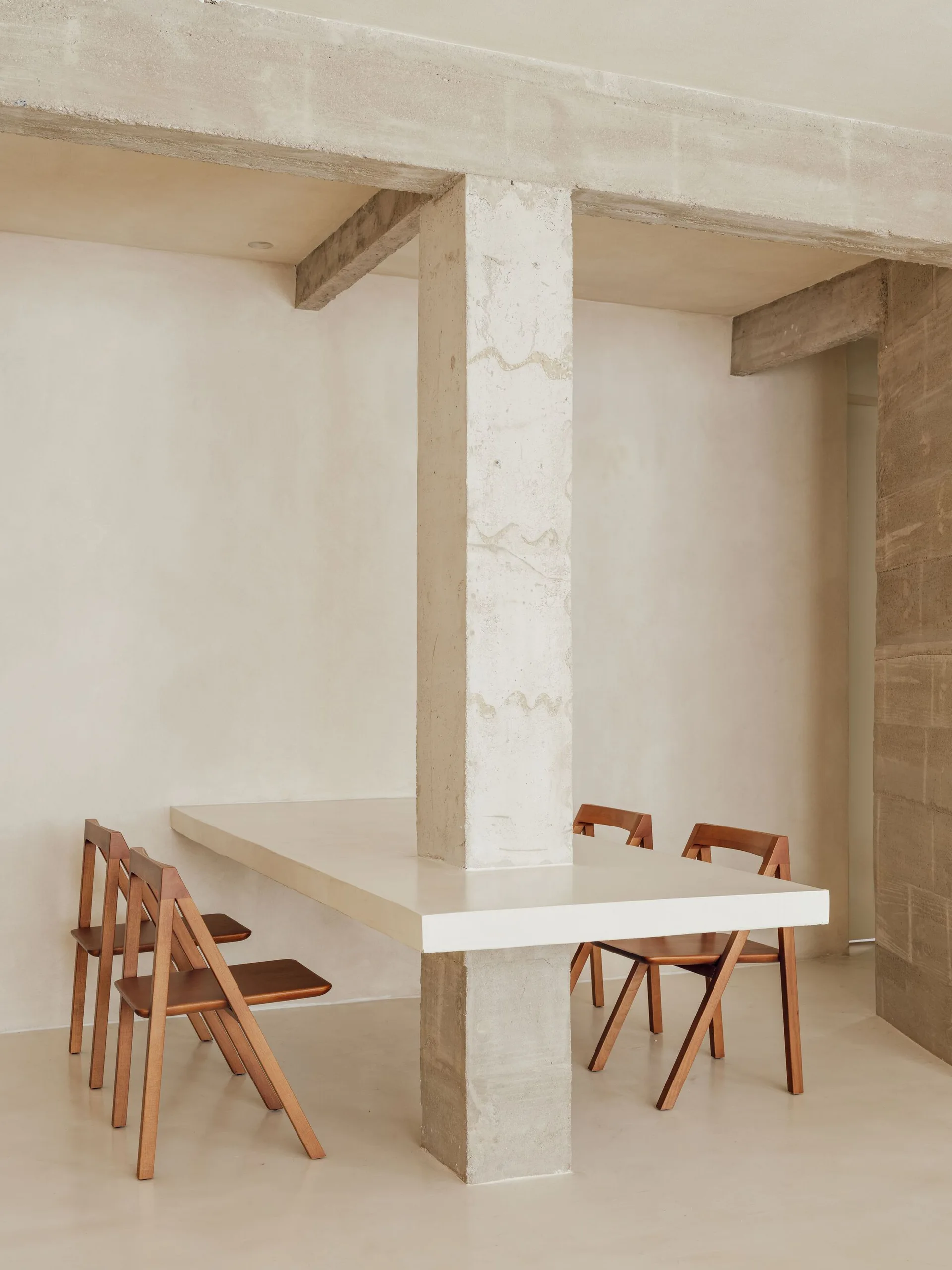 A white minimalist dining table is built directly into a central concrete column, accompanied by four wooden chairs in a neutral beige room.