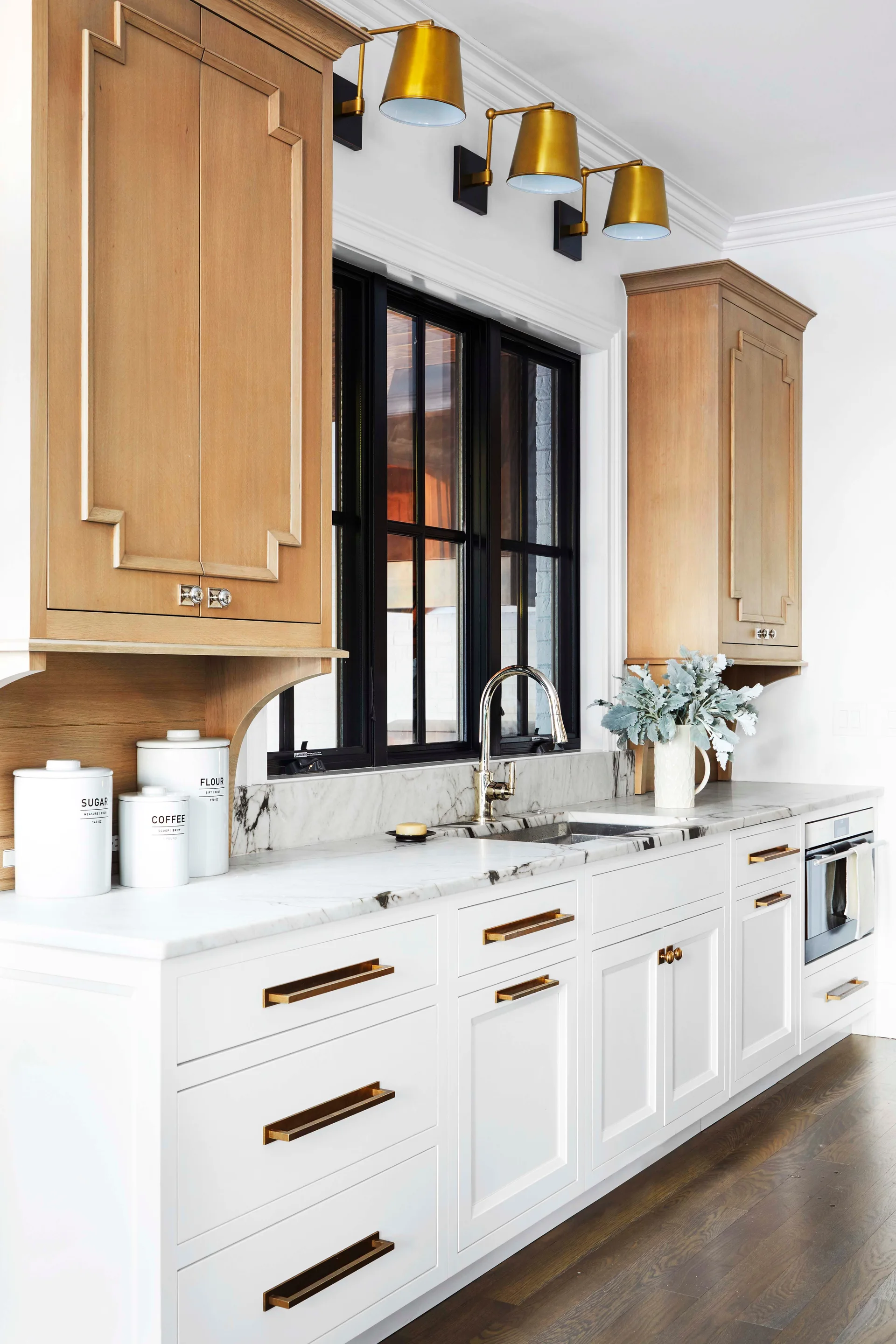 A kitchen counter features white base cabinets, light wood upper cabinets flanking a black window, marble countertops, and three brass wall sconces.